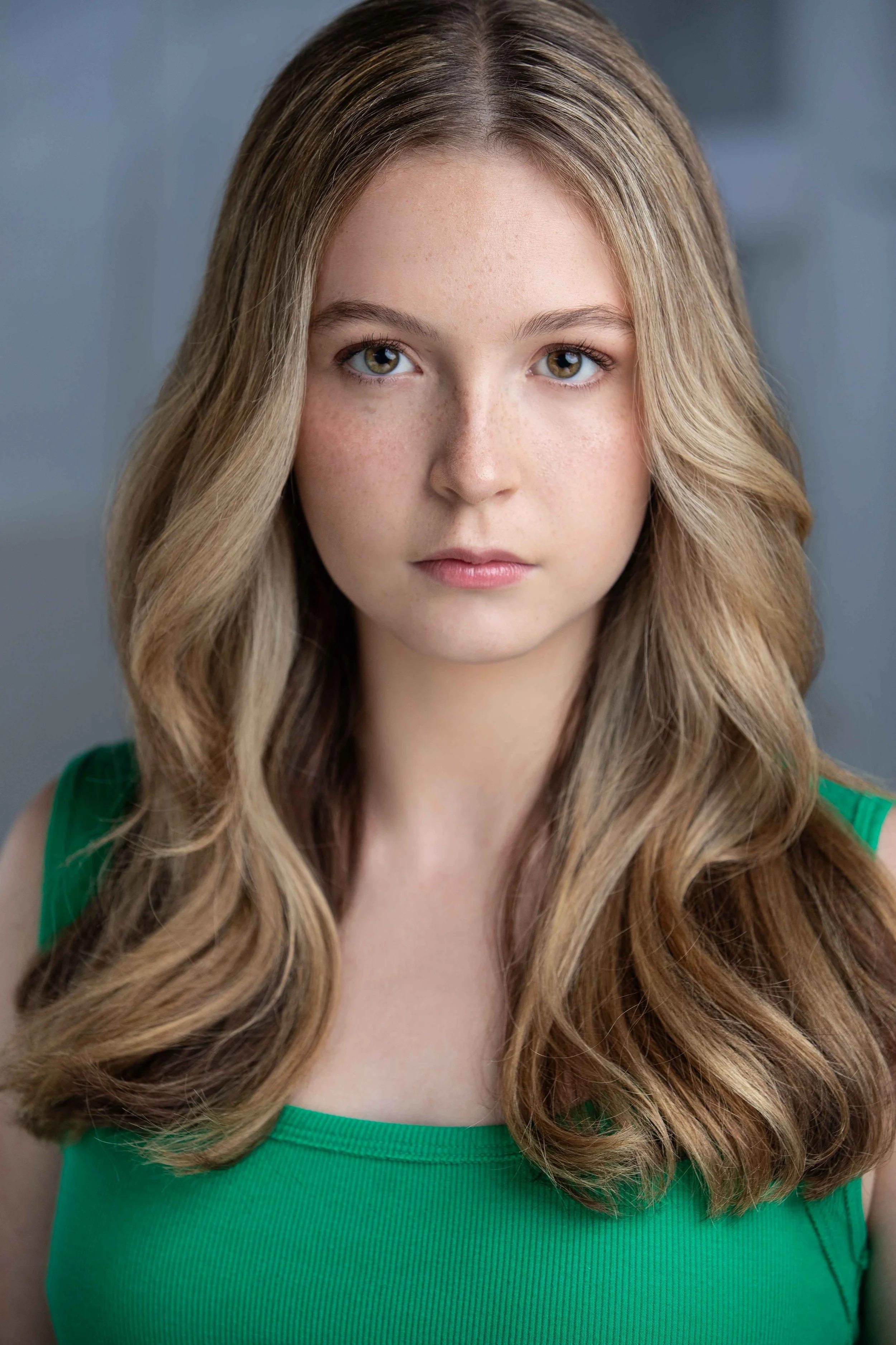 Close-up portrait of a young woman with long, wavy blonde hair, fair skin, and light eyes wearing a green top.