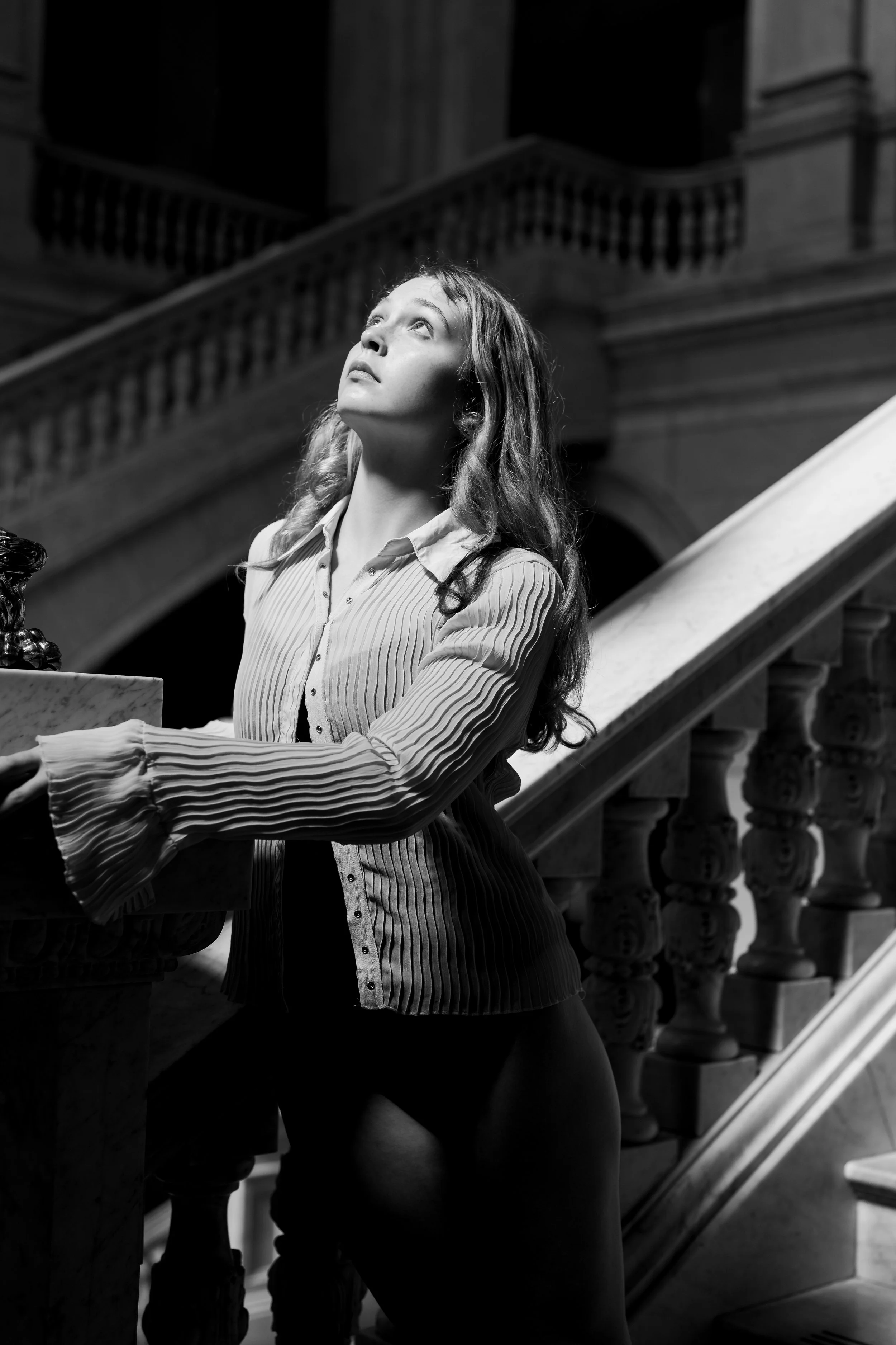 A young woman with wavy hair, wearing a striped blouse, stands on a staircase in a grand, historic building, looking upwards with a contemplative expression in black and white photo.