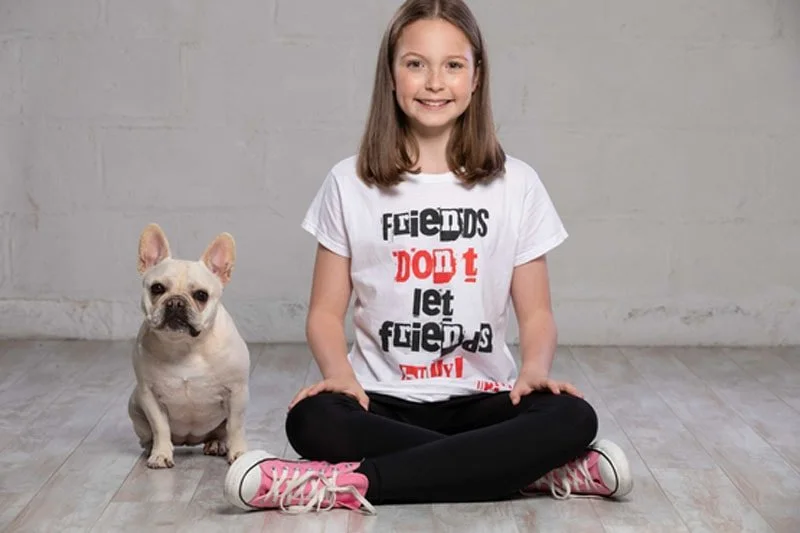 A girl sitting cross-legged on the floor next to a French Bulldog, both facing the camera, with a plain gray wall in the background.