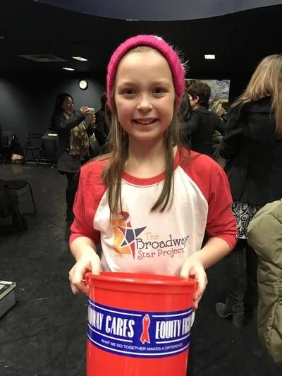 A young girl smiling and holding a red bucket at an indoor event. She is wearing a pink headband and a white and red shirt with a logo that reads "The Broadway Star Project." There are other people in the background.