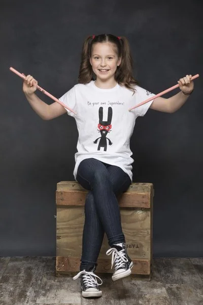 A young girl with long brown hair in pigtails sitting on a wooden crate, holding two pink pencils out to the sides, smiling at the camera, wearing a white T-shirt with a cartoon rabbit wearing a superhero mask and the text "Be your own Superhero!", d