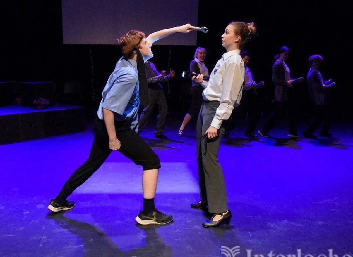 Two young people perform a theatrical scene on stage, with a group of girls in the background. One person holds a gun and is about to spray a liquid at the other person.