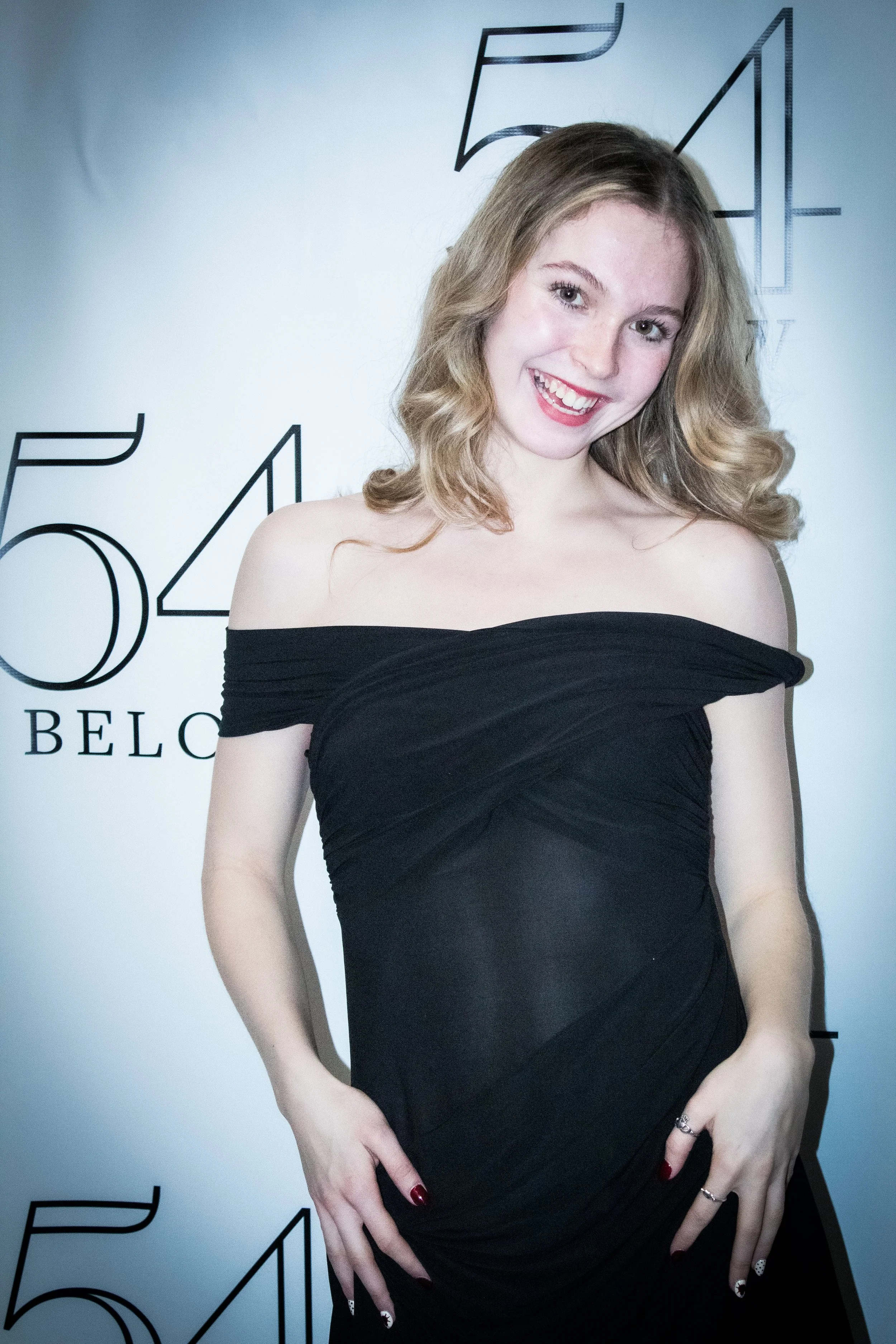 A woman with wavy blonde hair smiling and wearing an off-the-shoulder black dress, standing in front of a backdrop with numbers and partially visible text.