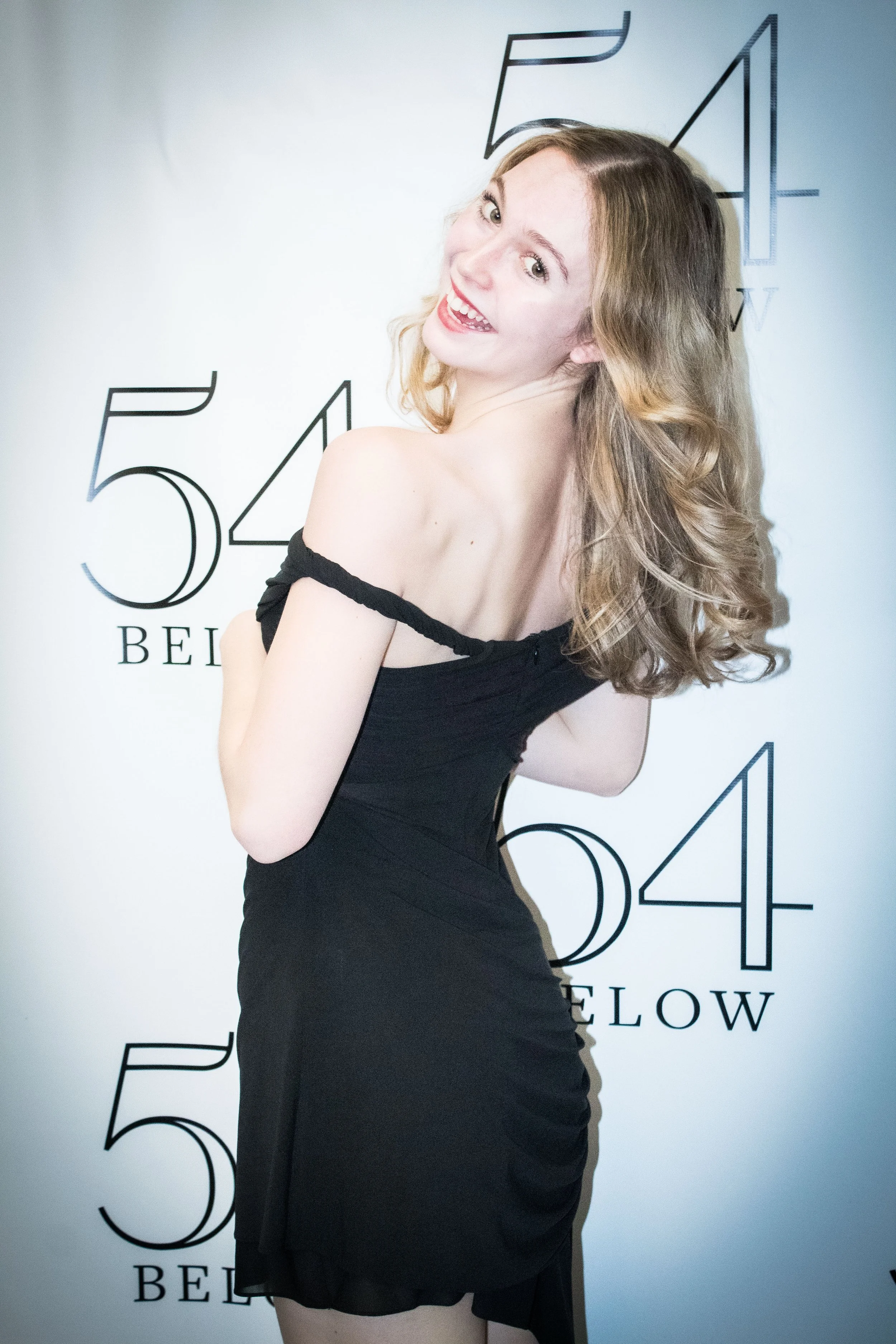 Woman in black dress smiling and posing on a step and repeat banner with the numbers 514 and the word Broadway.