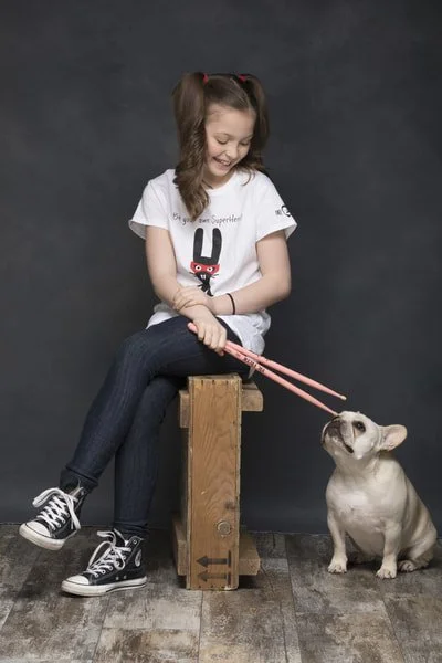 A young girl with brown hair in pigtails sitting on a wooden block, holding pink chopsticks and smiling at a French bulldog sitting on the floor.