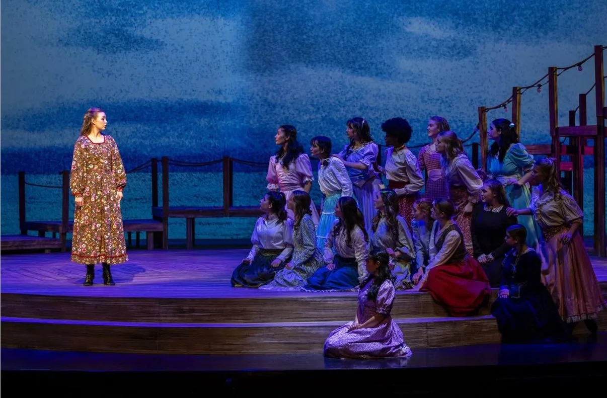 A group of children performing a play on stage, with one girl standing separate from the group, in a scene resembling a theatrical production.