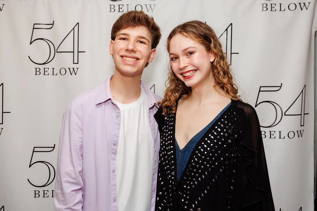 A young man and woman smiling and standing close together at a premiere event in front of a backdrop with '54 Below' written on it.