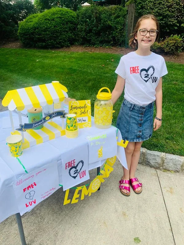 Young girl selling lemonade at a lemonade stand in a park or backyard with grass and bushes, wearing a white t-shirt and a denim skirt, smiling.