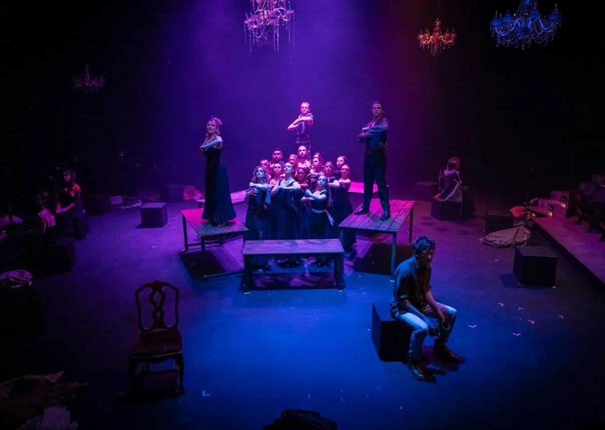 Theatrical stage with performers in dark costumes, some standing on platforms, under purple and blue lighting, with chandeliers hanging above and various props on stage.