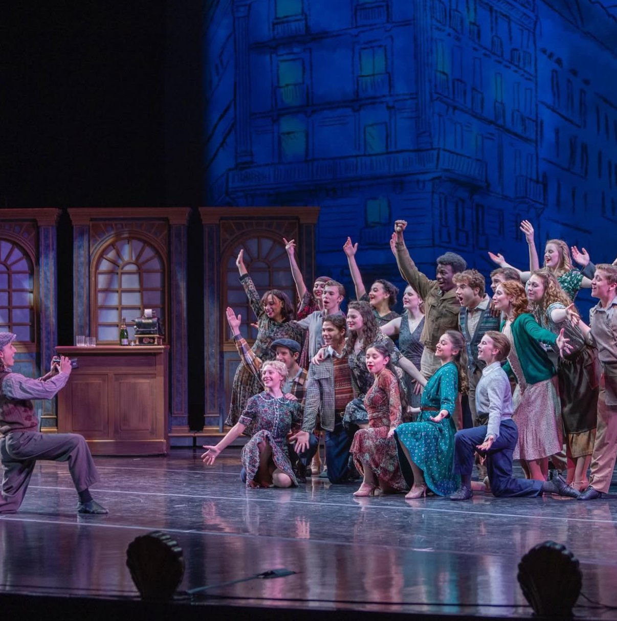 A group of performers on a stage, dressed in vintage clothing, participating in a theatrical production. They are smiling and engaged, with some kneeling and others standing, raising their hands in celebration or acknowledgment. The stage backdrop de