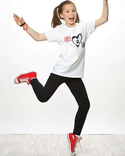 A young girl jumping in the air, smiling, with her arms raised. She wears a white t-shirt, black pants, and red sneakers.