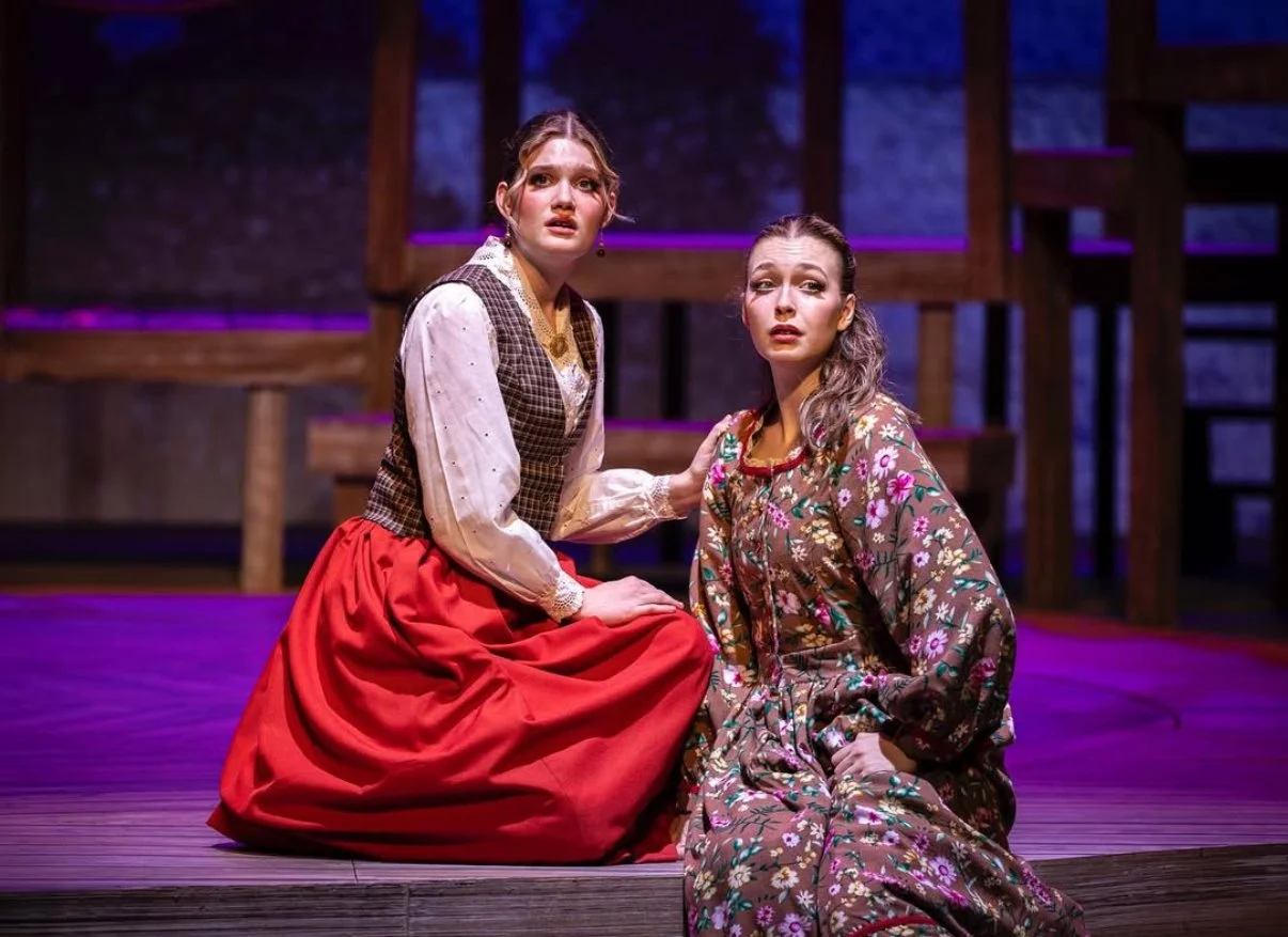 Two women in period costumes performing on stage, one kneeling and the other sitting, with a wooden set background and stage lighting.