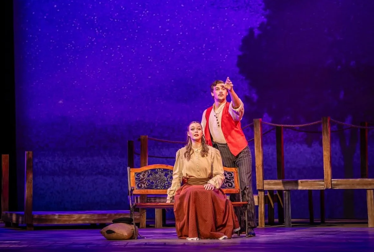 The scene is part of a theatrical play with two actors on stage, a woman sitting on a wooden bench and a man standing beside her pointing upwards. The background depicts a nighttime sky with stars. Wooden fencing and a bag are also visible on the sta