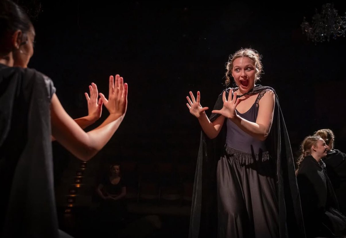 Actors on stage performing in a theatrical play, with one woman in a dark dress and cloak reacting dramatically to two other actors' gestures.