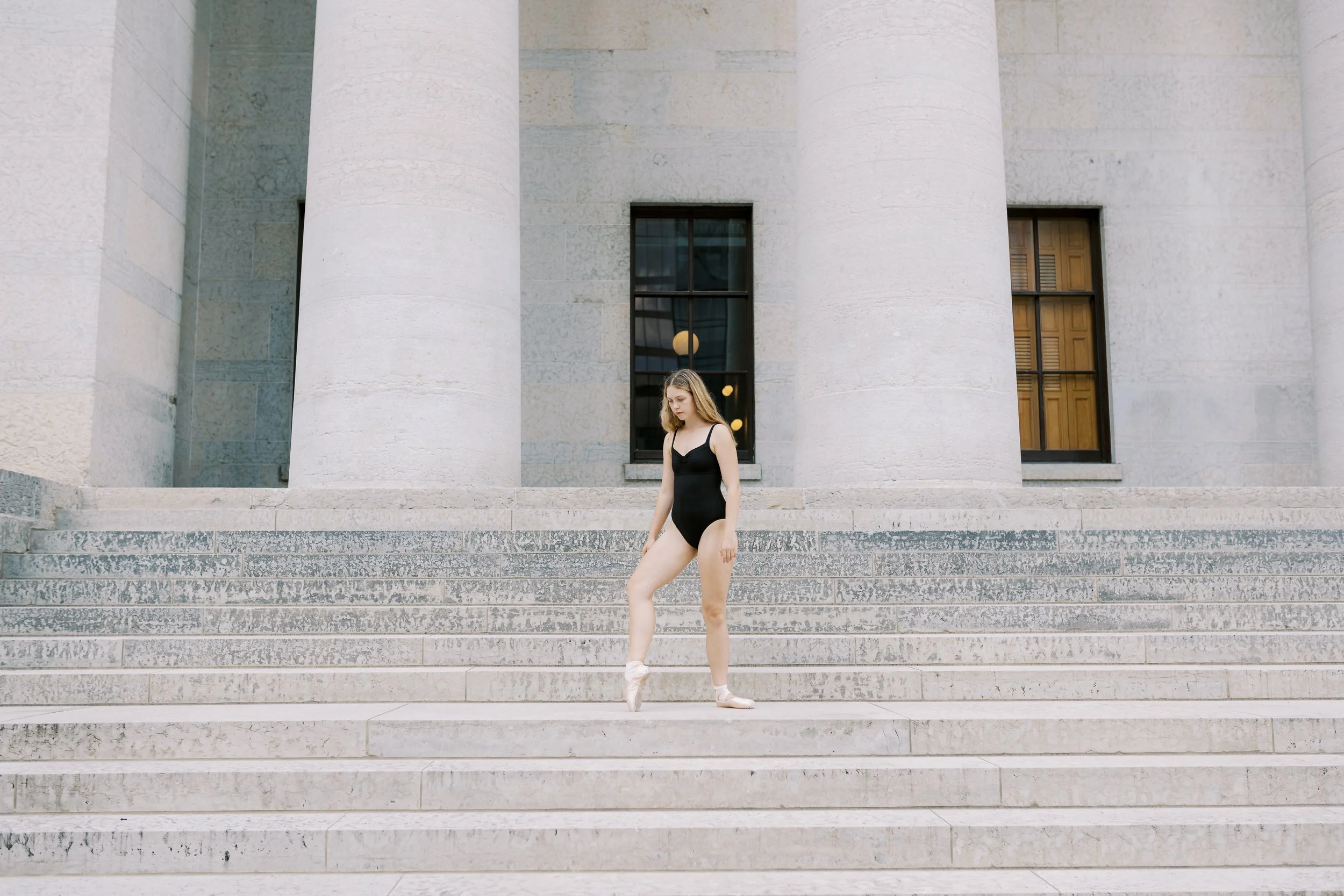 A young woman dressed as a ballet dancer in a black leotard, pink ballet shoes, standing on the steps of a large classical building with tall columns and rectangular windows.