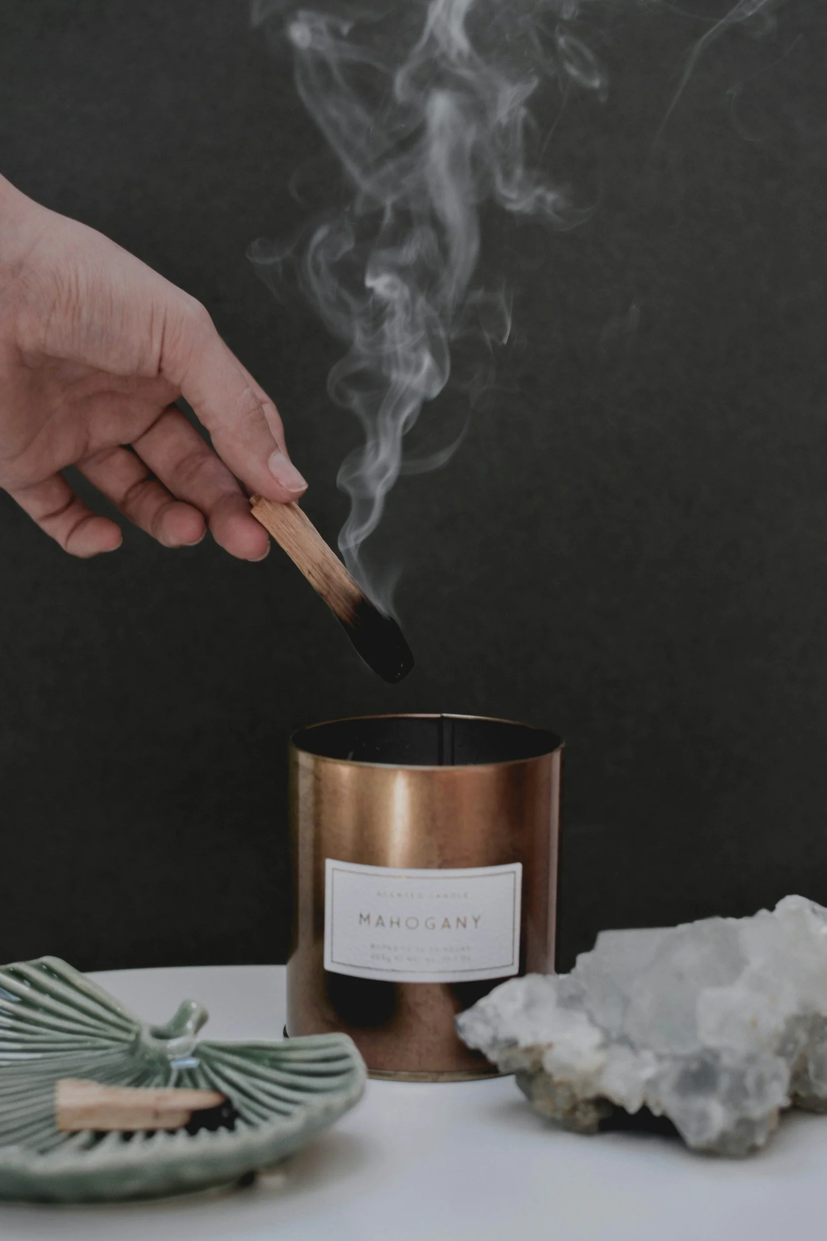 A hand holding a lit incense stick emitting smoke over a copper-colored candle holder labeled "Mahogany" with a white rocky object and a decorative ceramic bowl nearby.