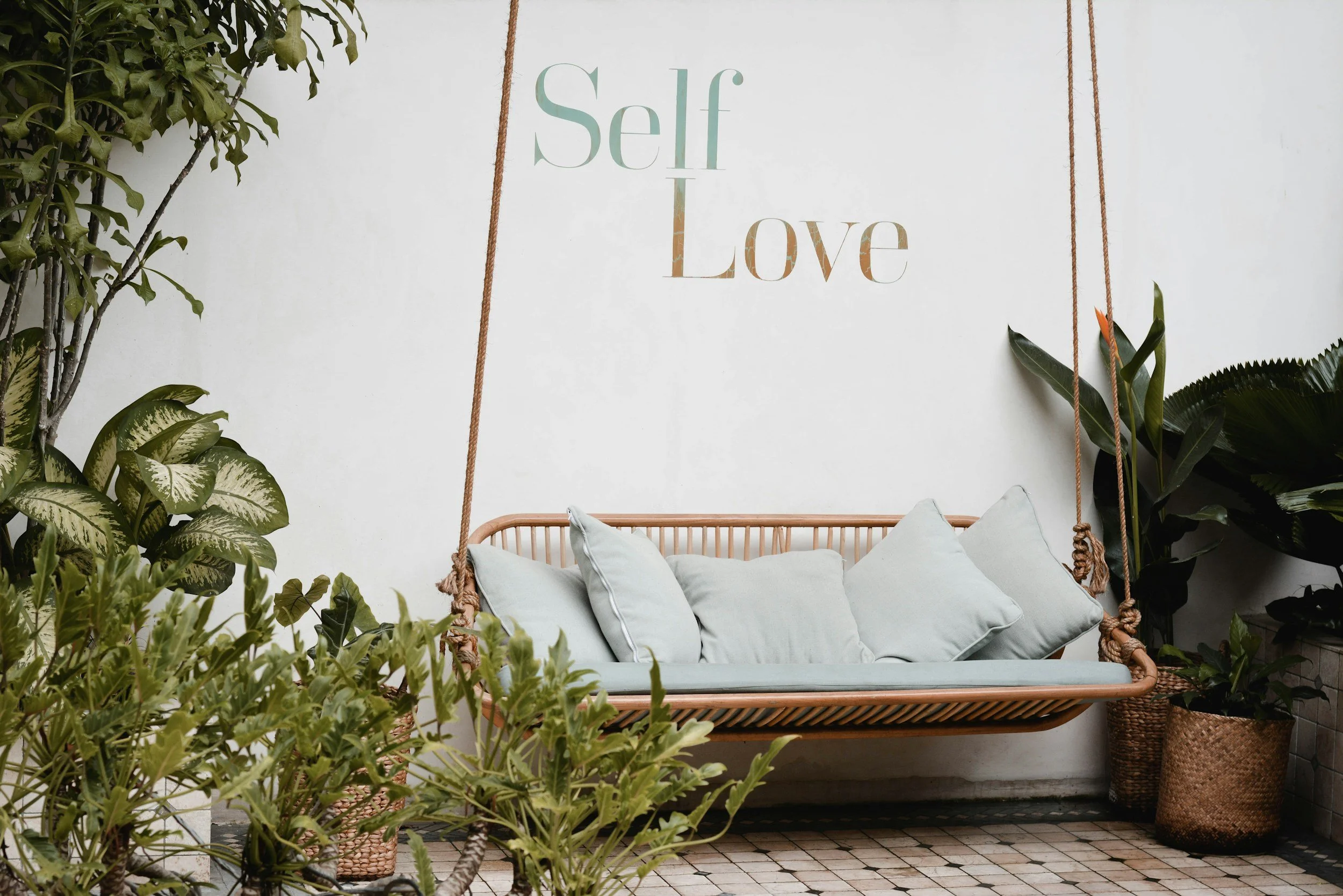 A white hanging swing with cushions, surrounded by lush green plants, with a wall sign that reads 'Self Love' in large pastel-colored letters.
