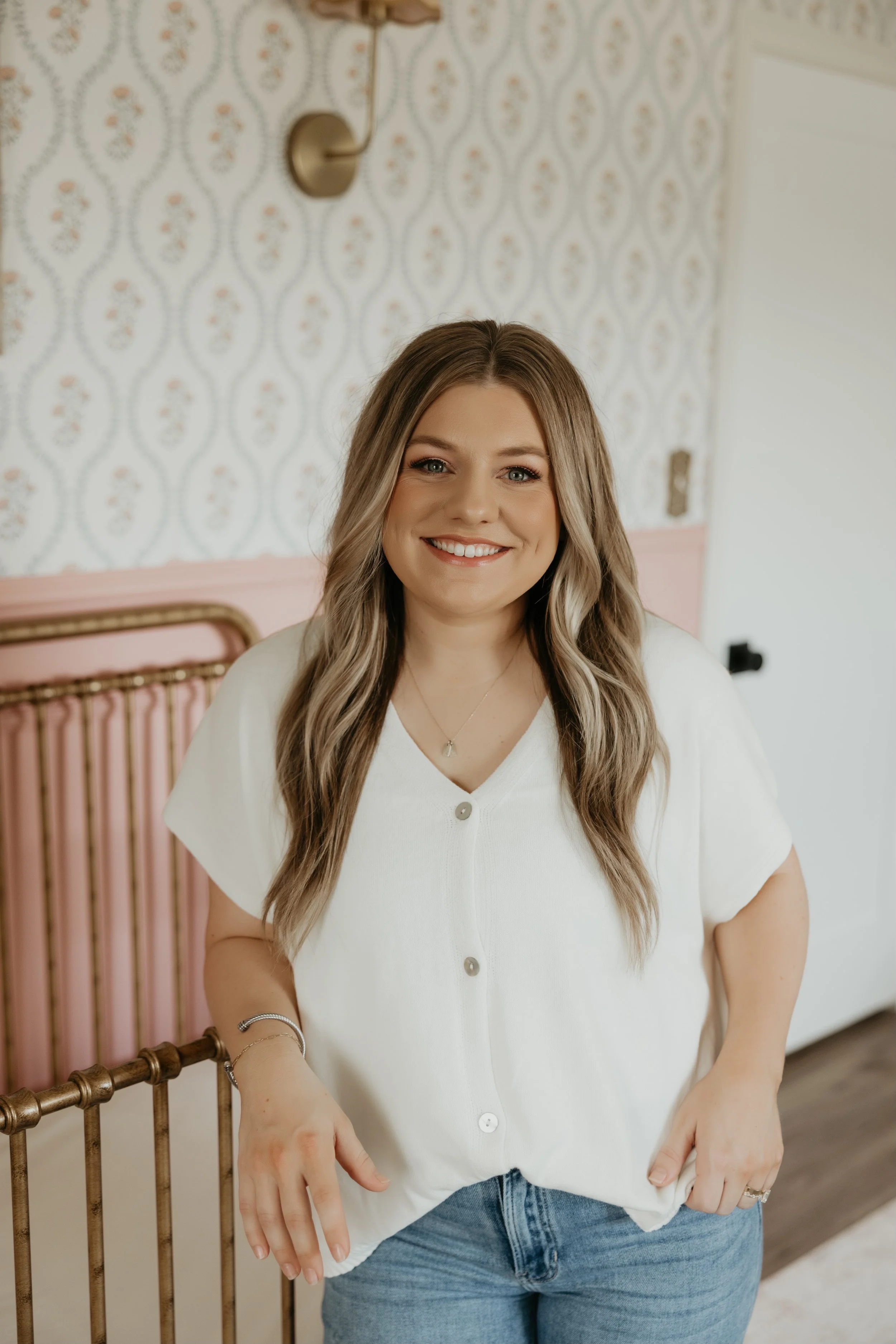 A smiling young woman with long wavy hair, wearing a white button-up shirt and light blue jeans, standing next to a vintage pink crib in a room with patterned wallpaper and a brass wall lamp.