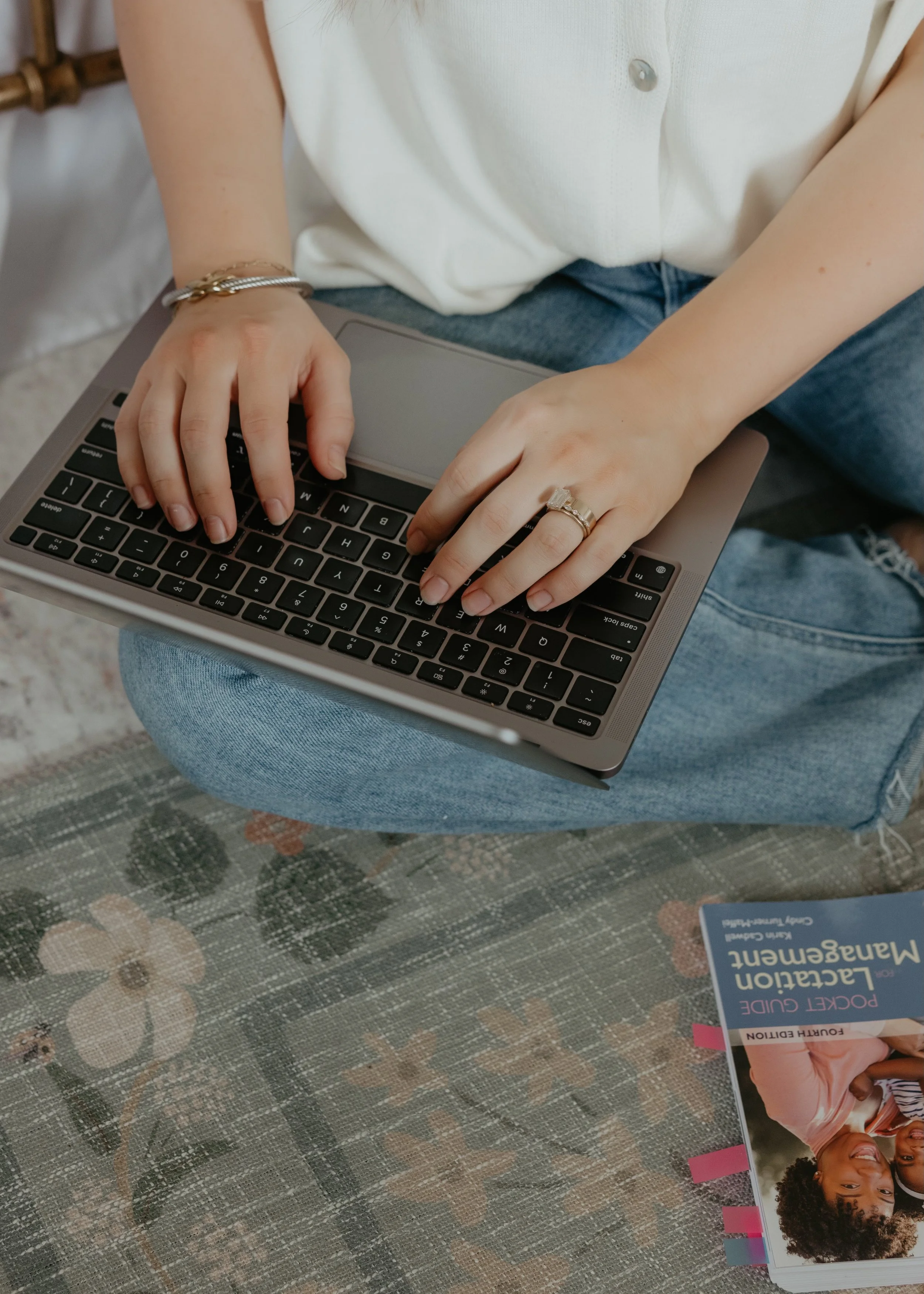 Person typing on a laptop keyboard while sitting cross-legged on a floral-patterned rug, with a booklet titled 'Education for Mental Wellbeing' nearby.