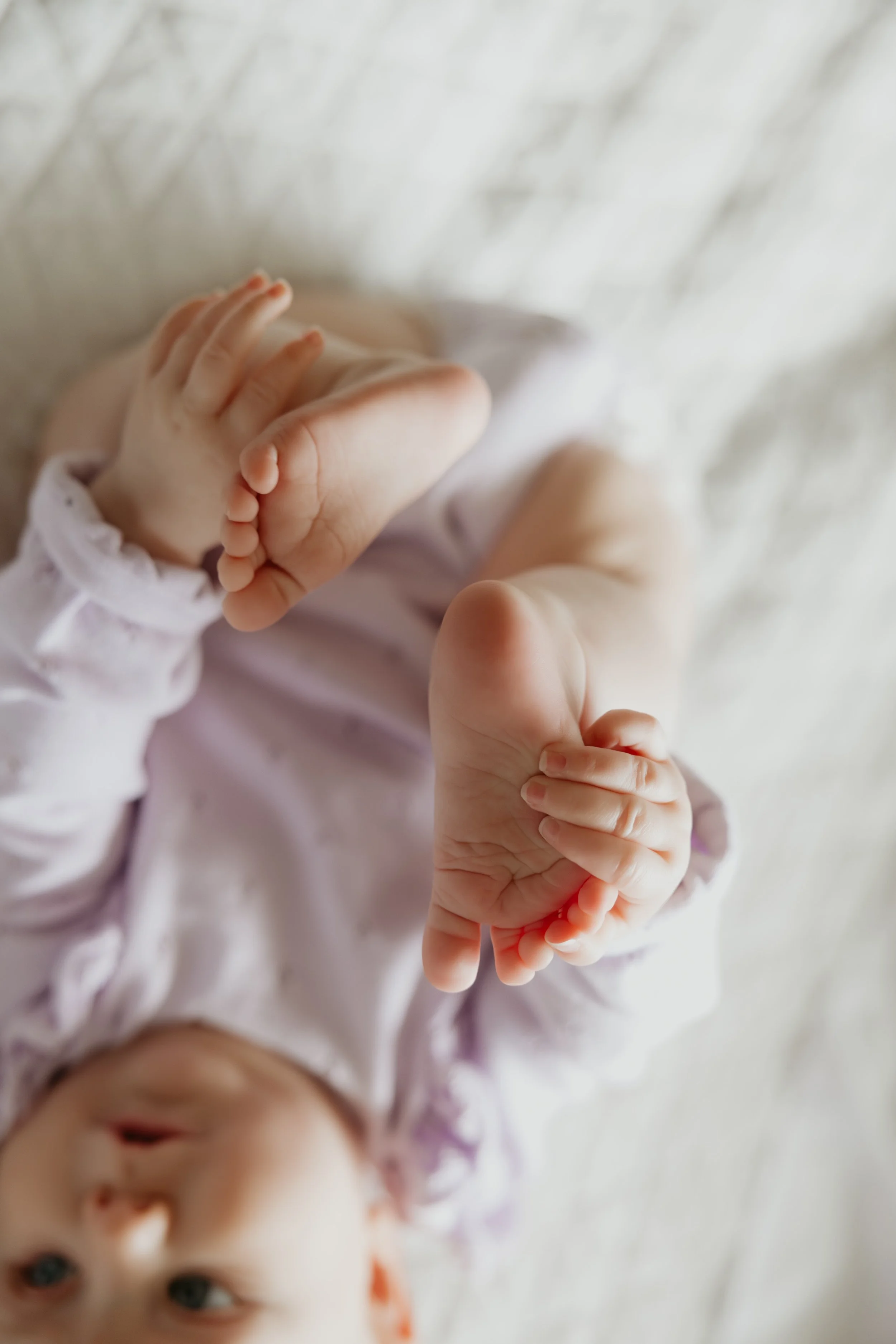 A close-up of a baby's tiny feet, lying on a soft, light-colored surface, with the baby's face slightly blurred in the background.