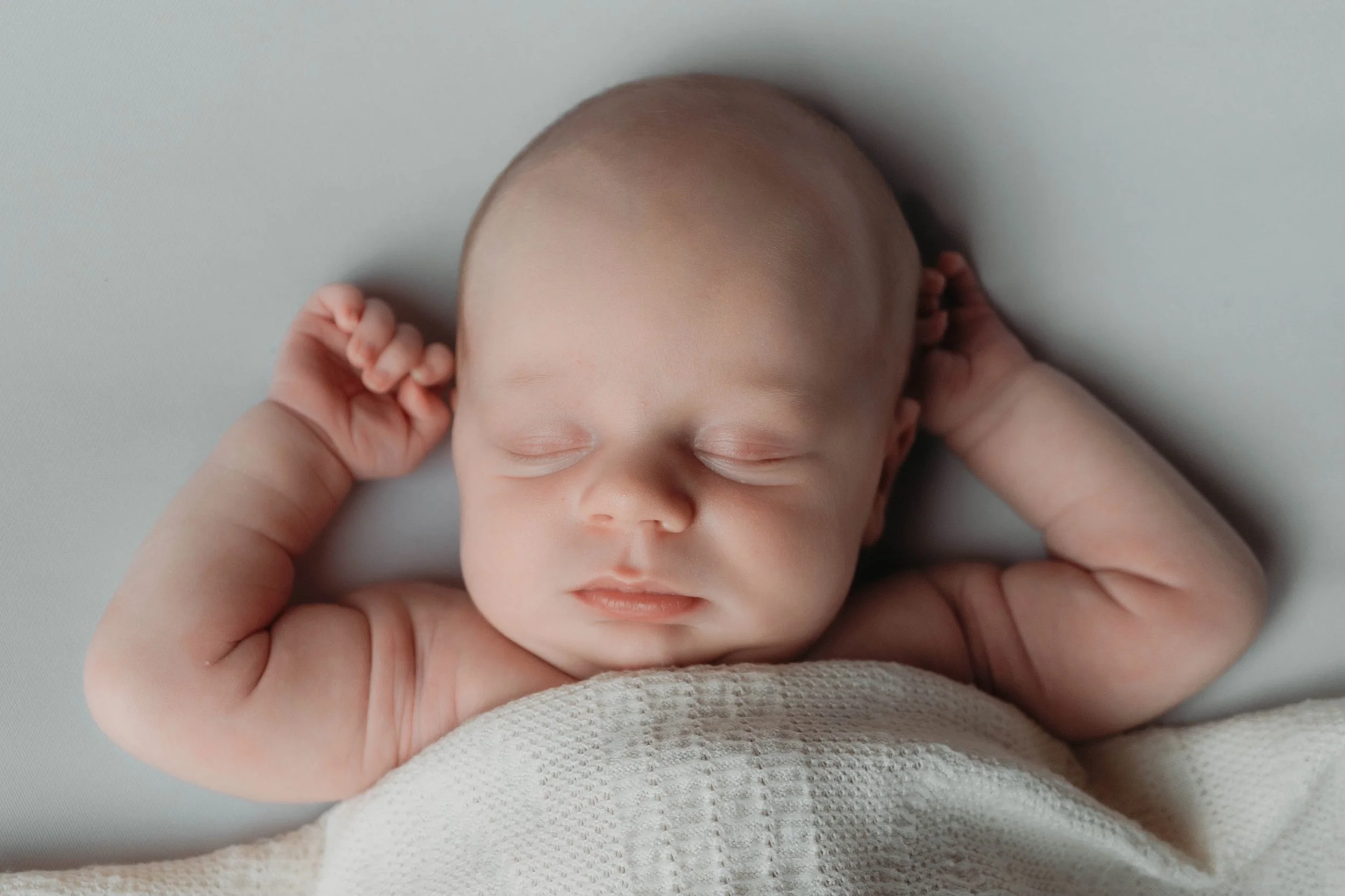 A sleeping baby with arms raised, peacefully resting on a soft surface, wrapped in a light-colored blanket.