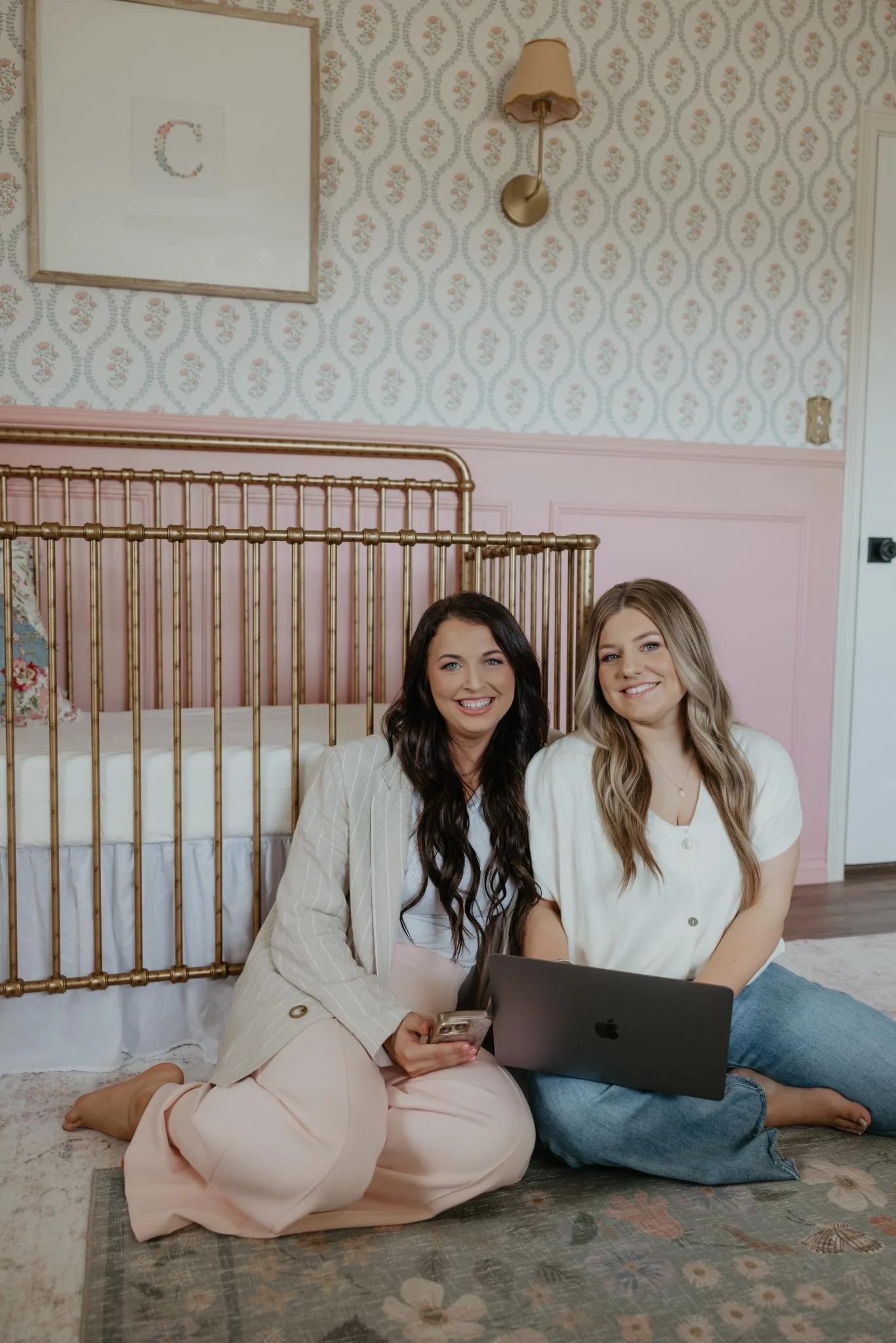 Two women sitting on a rug in a pink-themed room with wallpaper, with a gold crib behind them. One woman holds a smartphone and the other a laptop, both smiling.