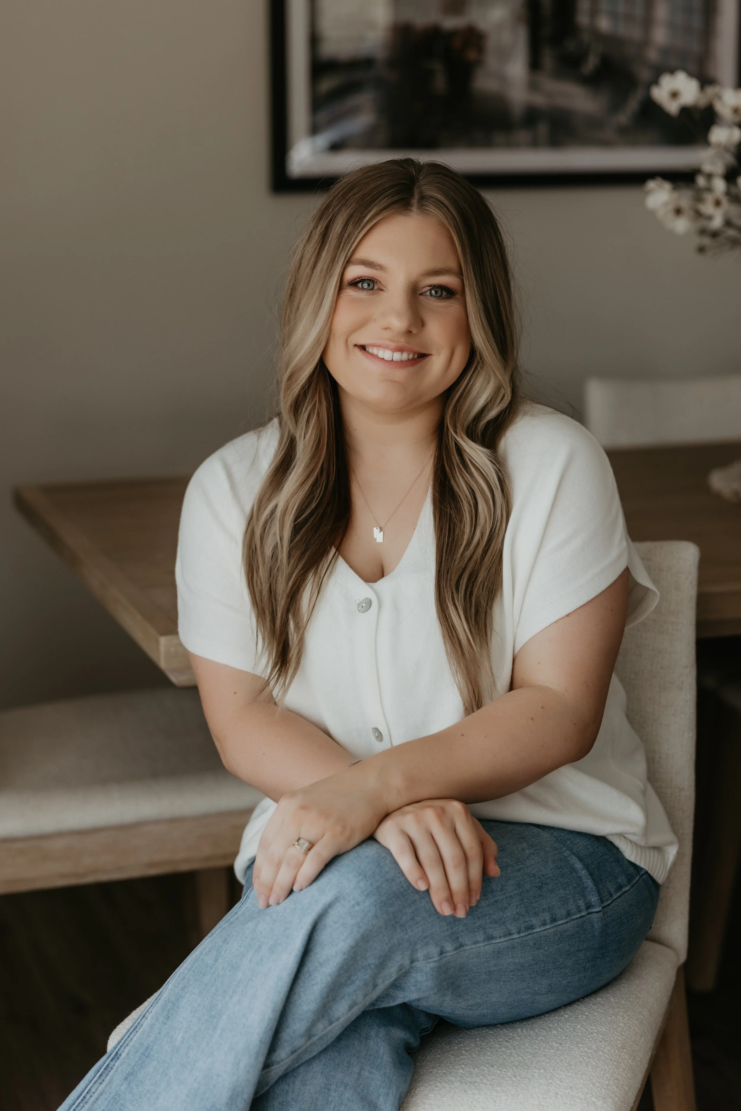 A woman with long wavy blonde hair, wearing a white button-up shirt and blue jeans, smiling and sitting in a beige chair in a cozy indoor setting with a wooden table and framed picture in the background.