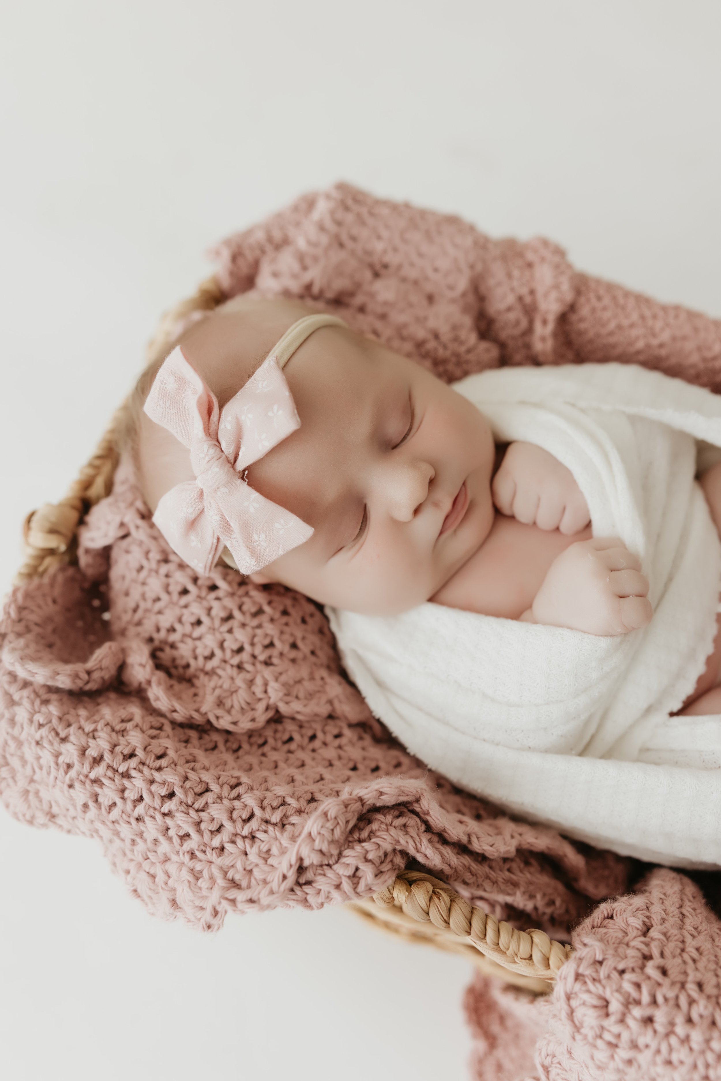 A sleeping baby with a pink polka dot bow headband, wrapped in a white blanket, lying on a pink knitted blanket inside a straw basket.