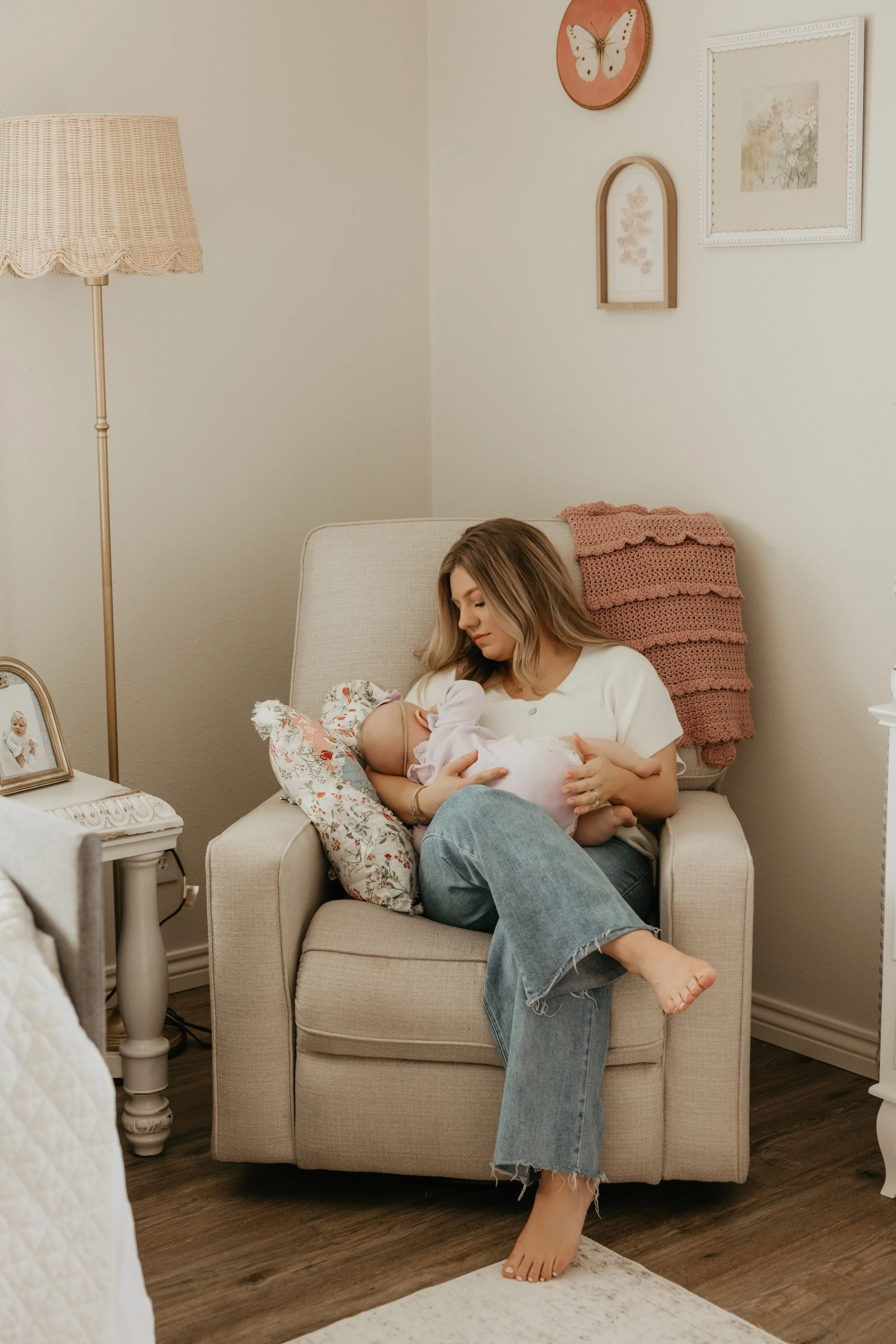 A woman sitting on a beige armchair breastfeeding a baby in a cozy, well-decorated room with soft pastel walls, framed artwork, and a pink knitted blanket over the back of the chair.