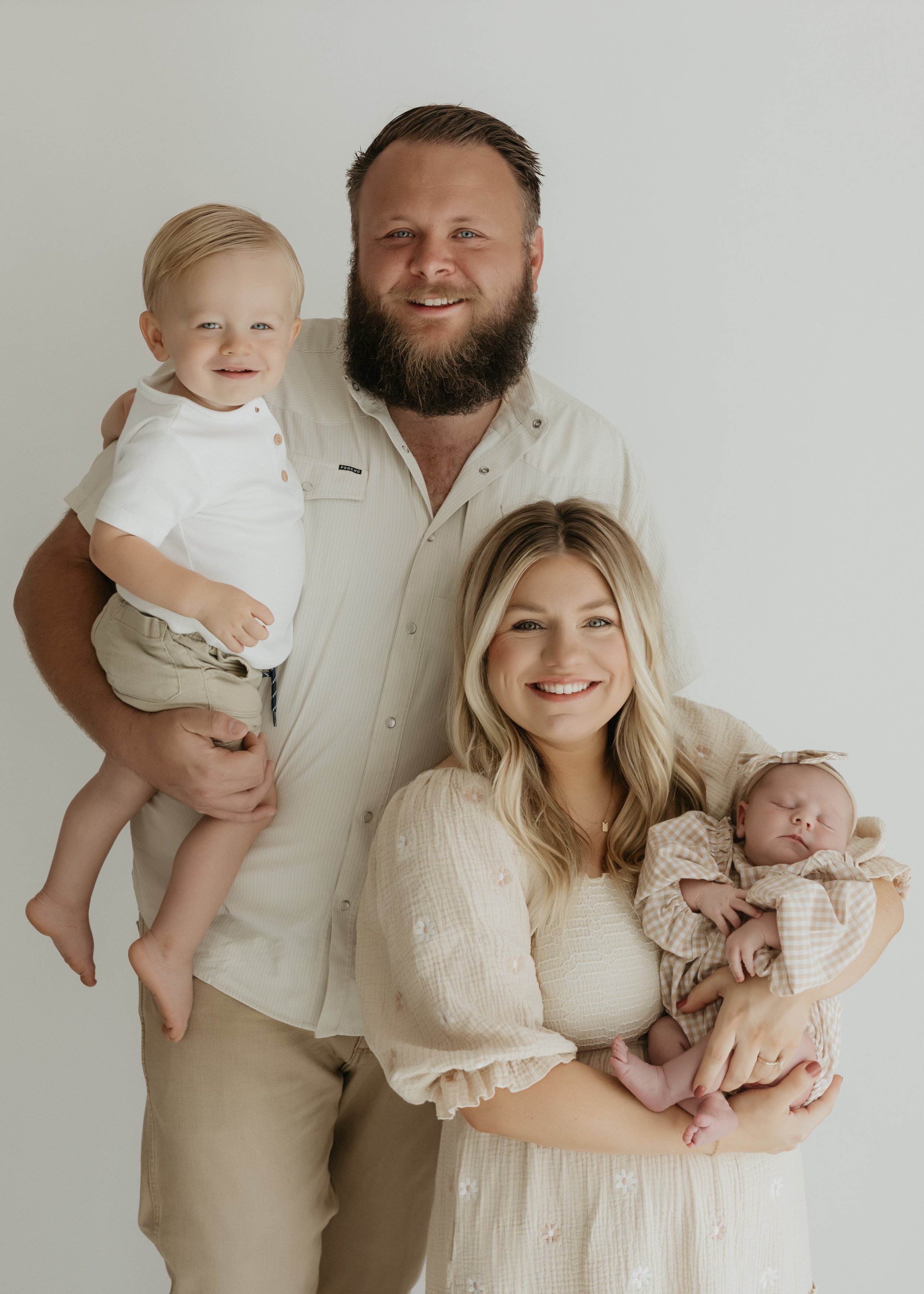 A family of four smiling, with a bearded man holding a young boy, a woman holding a sleeping baby, standing against a plain light background.