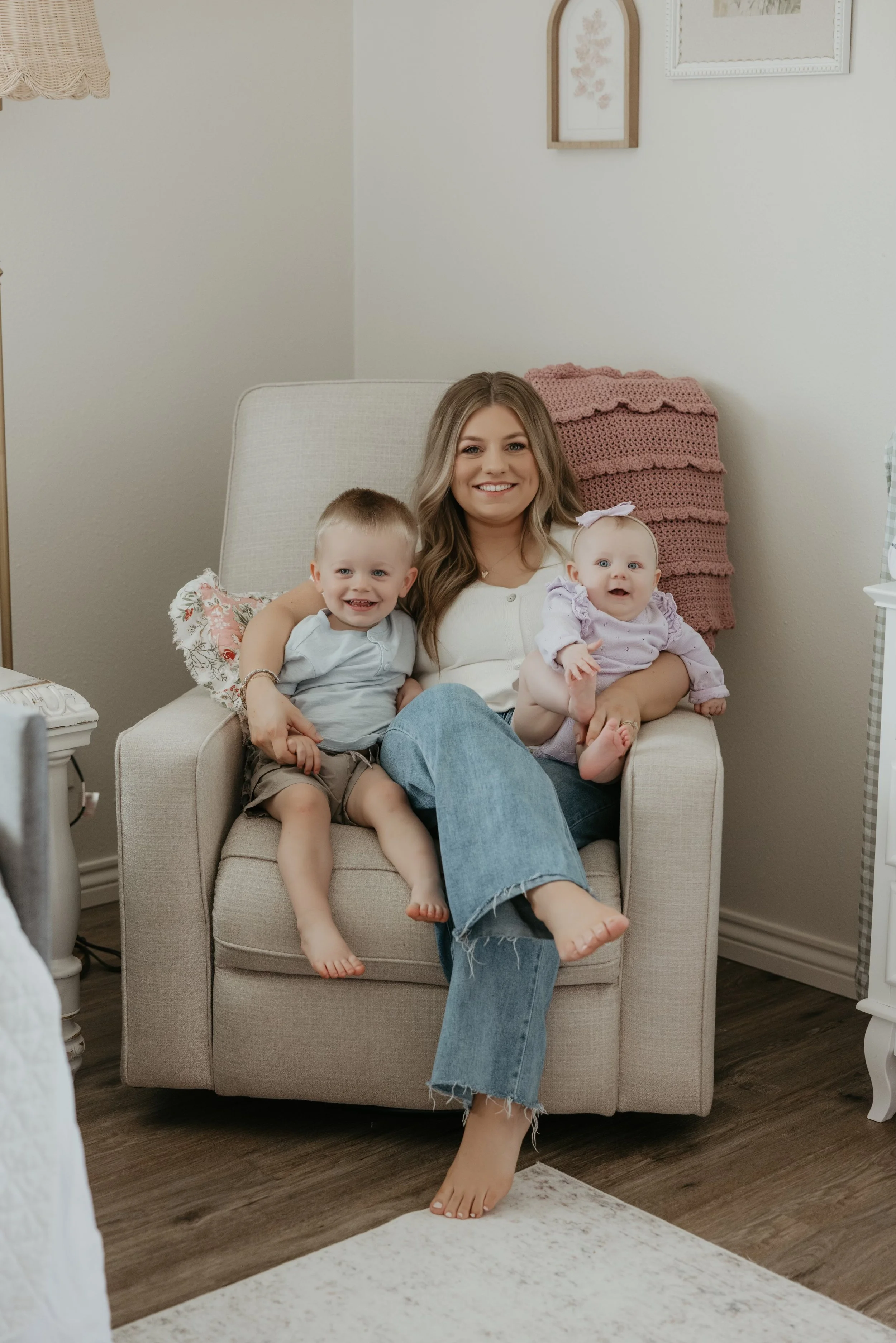 A woman with long wavy hair sitting in an armchair, holding a young boy on her left knee and a baby girl on her right arm, in a cozy room with decor and wooden flooring.