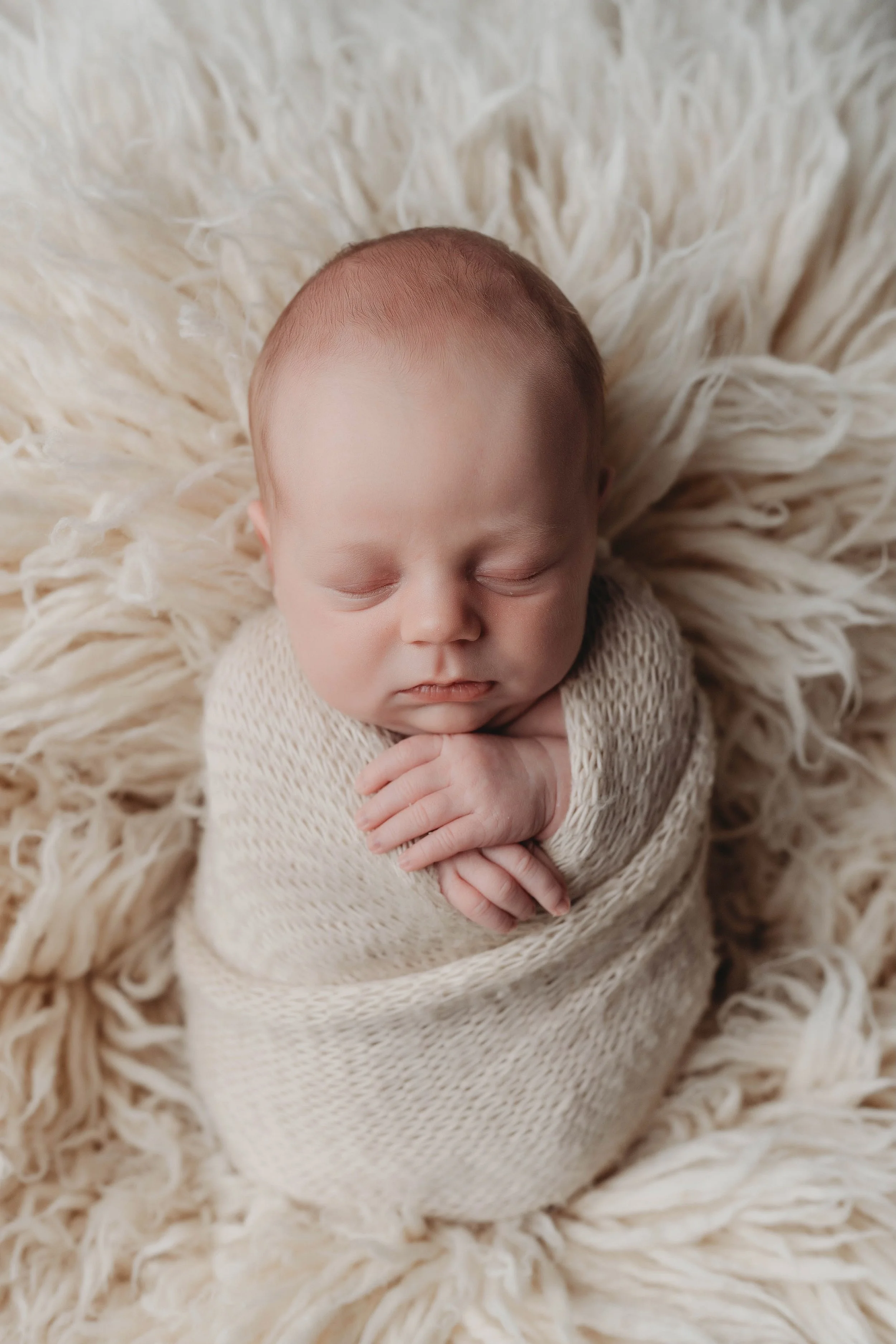 A sleeping newborn baby swaddled in a beige knit blanket, lying on a fluffy cream-colored blanket.