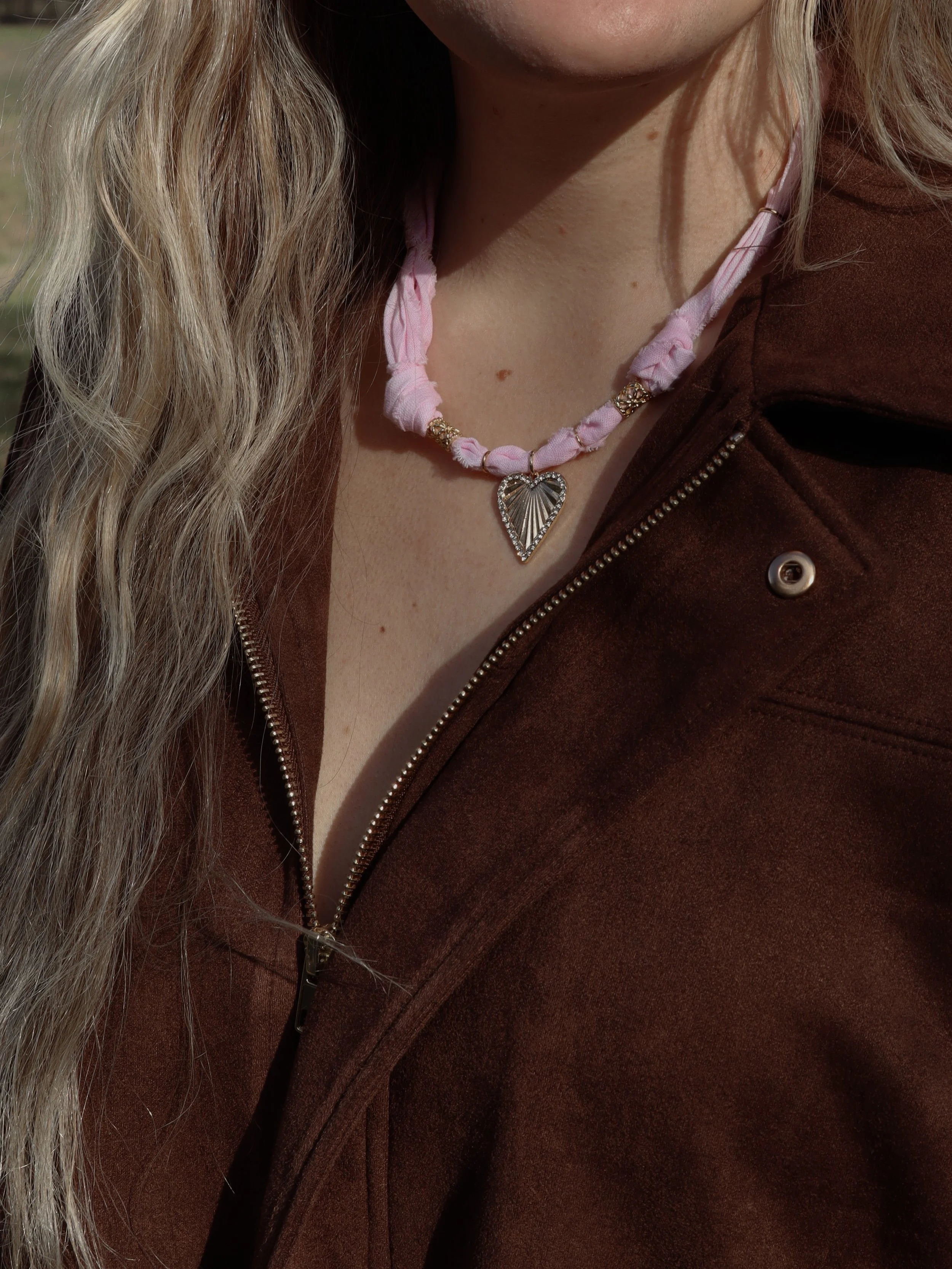 A woman wearing a pink fabric necklace with a heart pendant and a brown zip-up jacket.