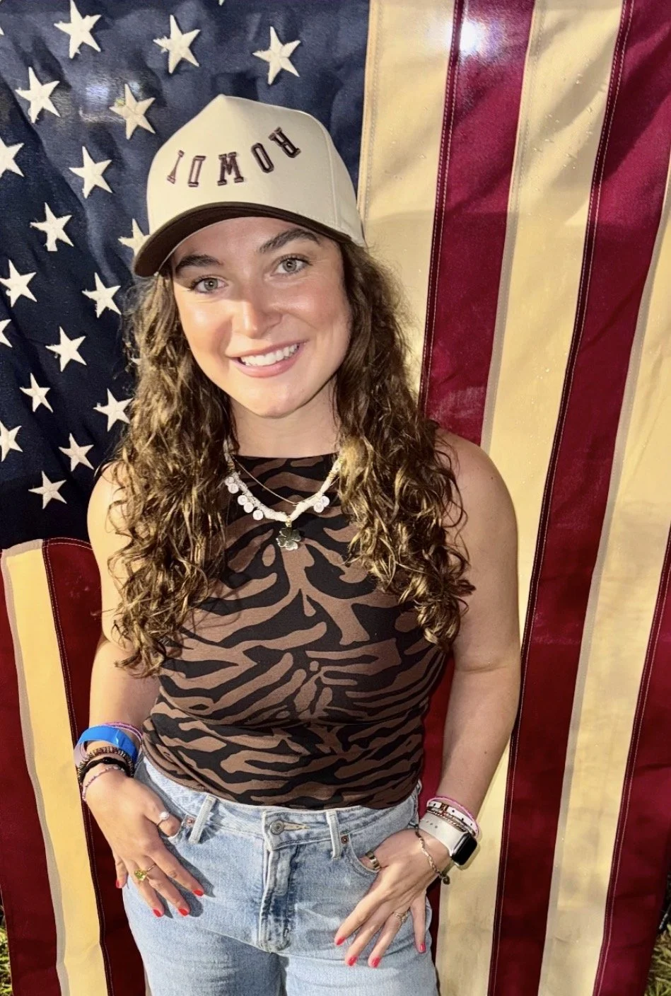 Young woman smiling, wearing a baseball cap with the word 'ROWDY,' zebra print sleeveless top, light blue jeans, standing in front of a large American flag.