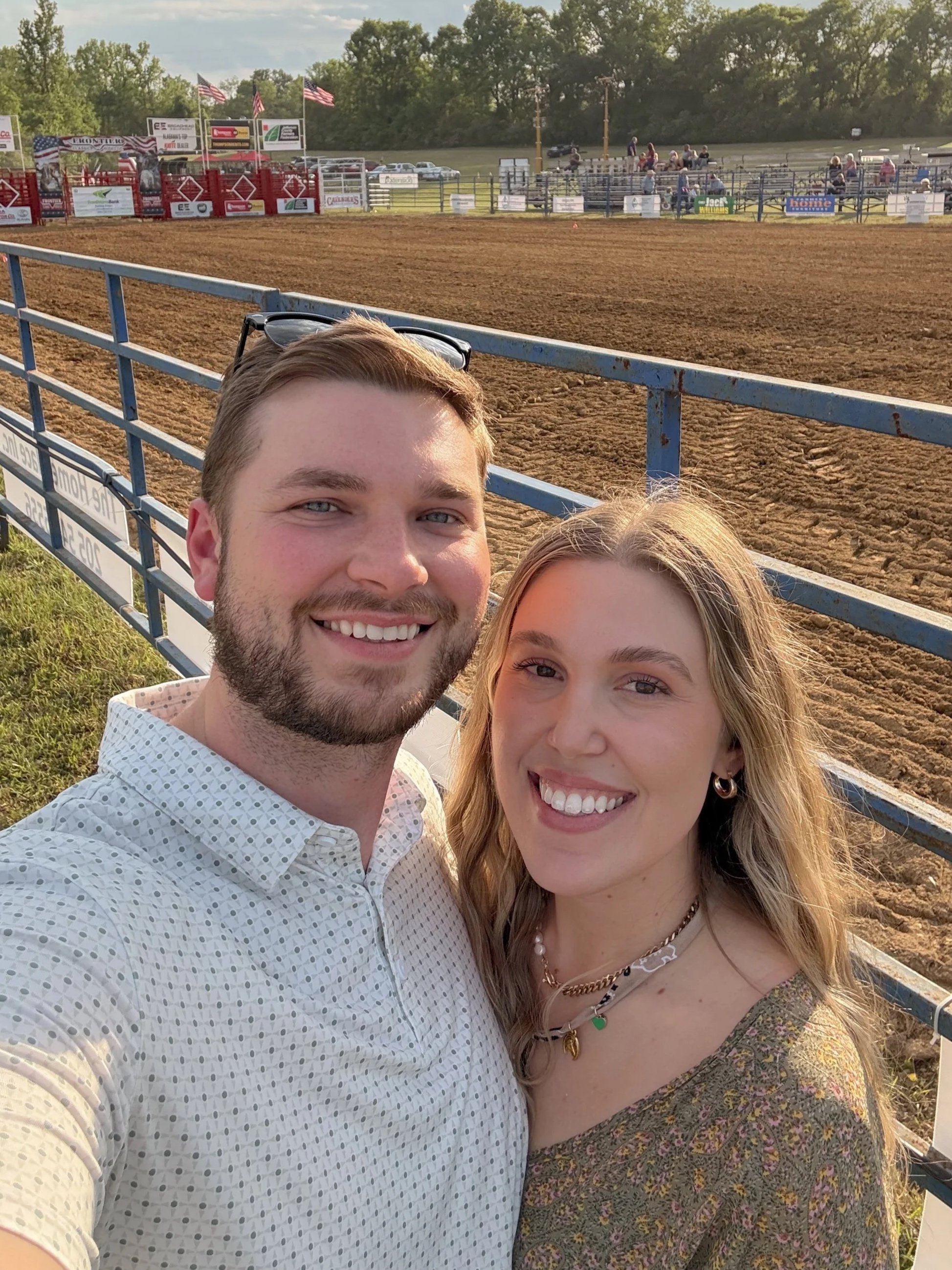 A smiling man and woman taking a selfie at a dirt rodeo arena with bleachers and American flags in the background.