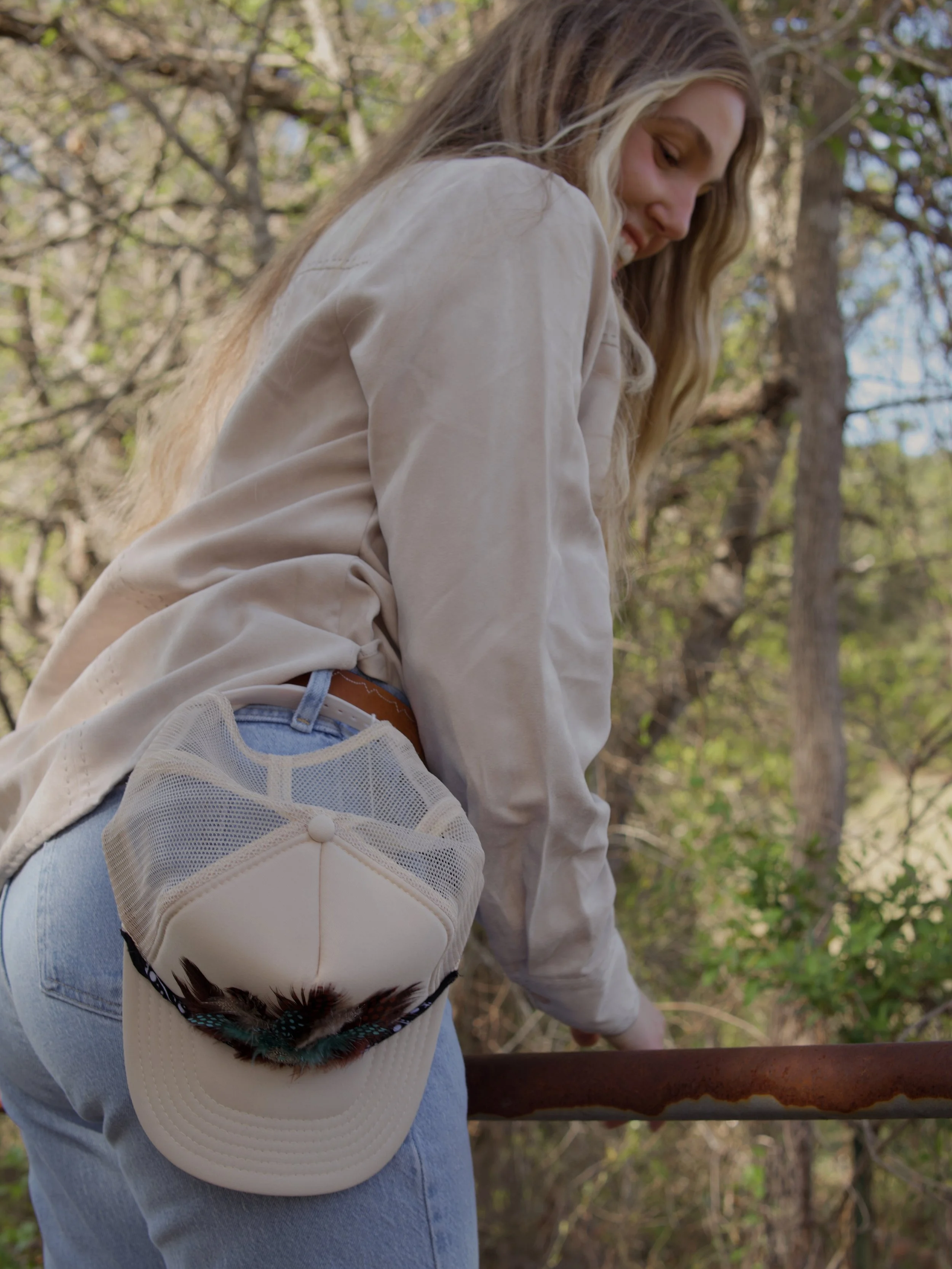 A young woman with long blonde hair wearing a beige jacket and blue jeans leaning over a rusty railing outdoors in a wooded area.