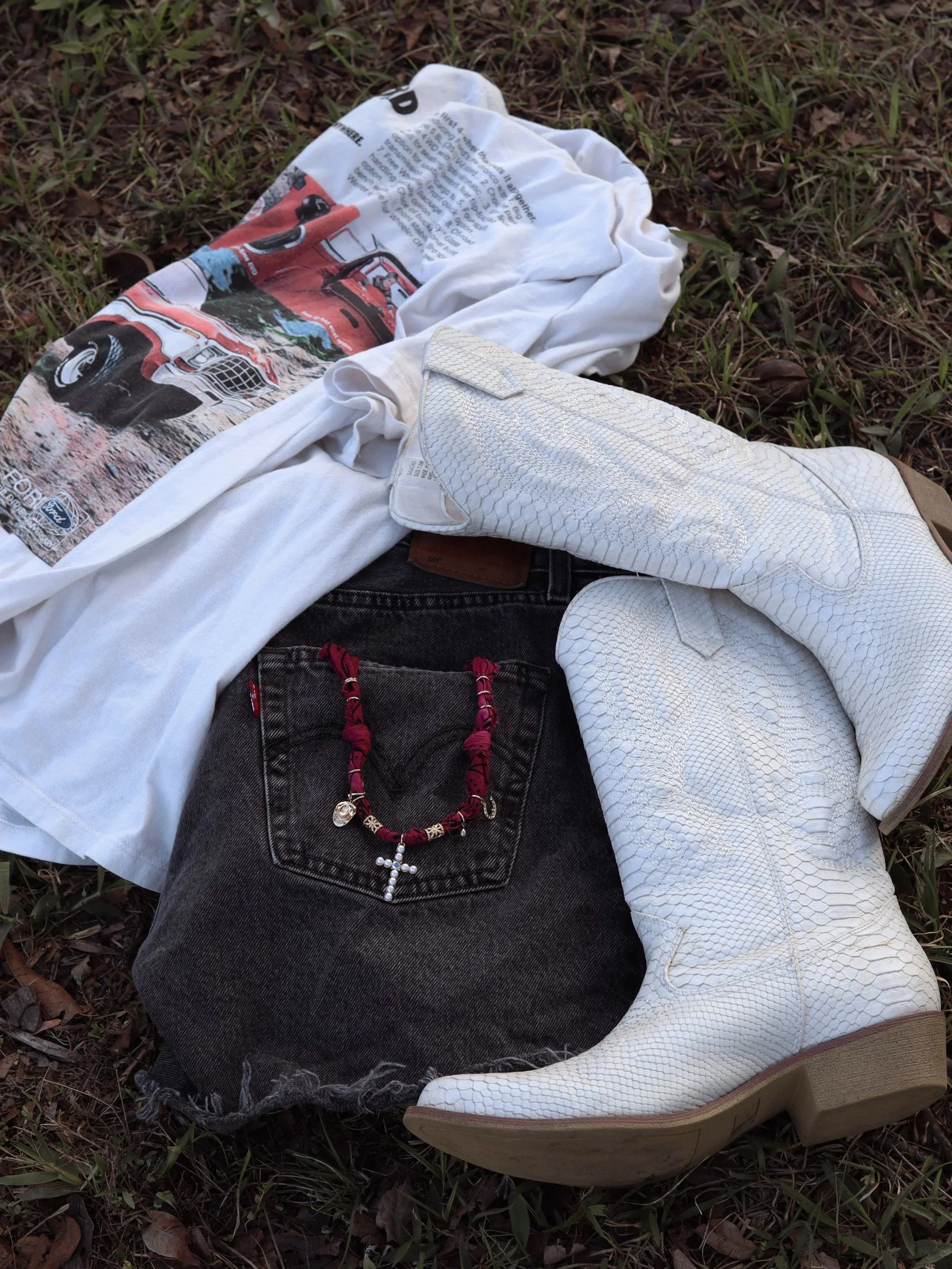 Clothing and accessories laid on the ground, including a white graphic T-shirt, black denim shorts with a red necklace and cross charm, and white snakeskin cowboy boots.