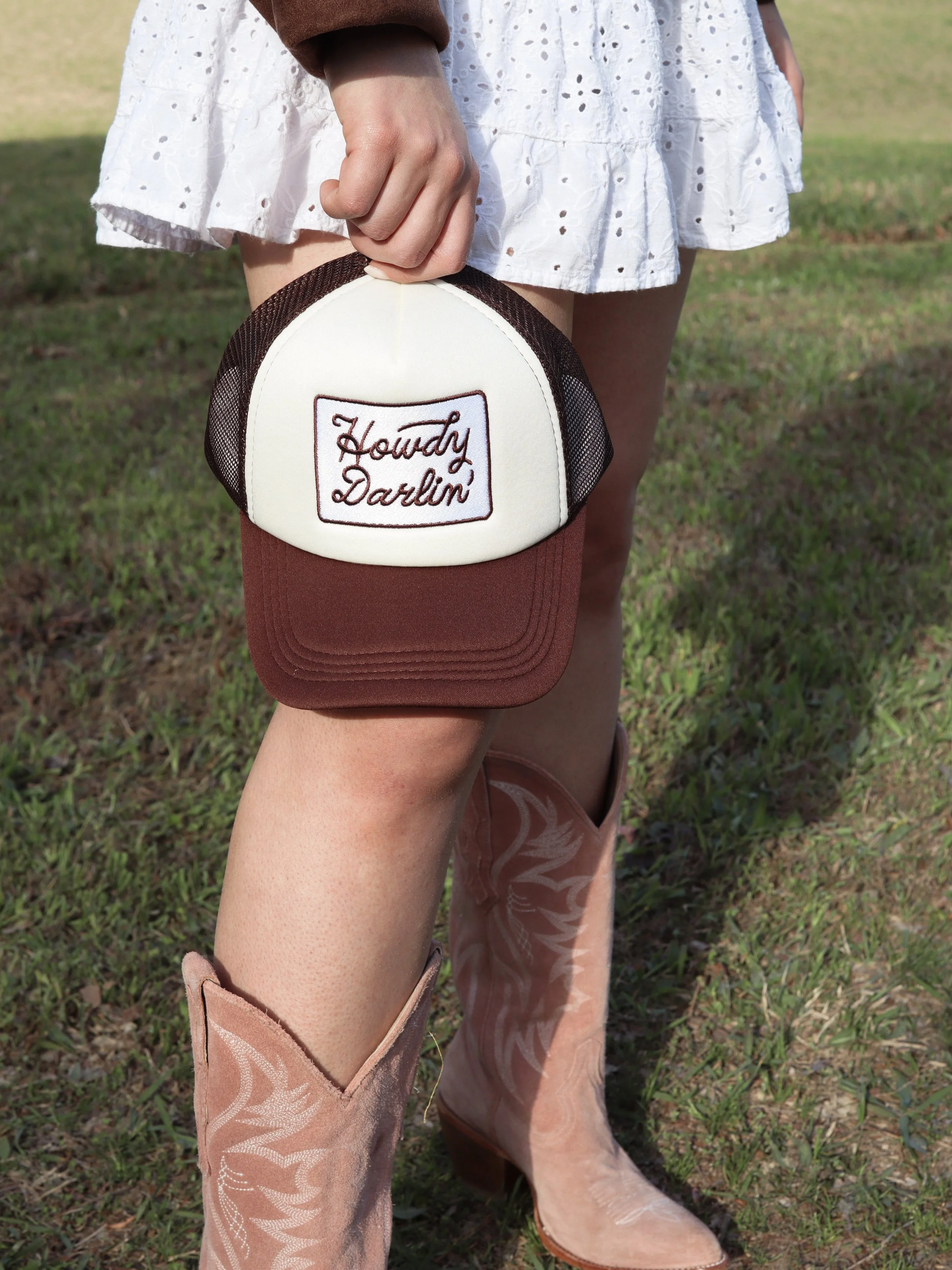 Person wearing cowboy boots and a white skirt, holding a trucker hat with 'Howdy Darlin' embroidered on the front in a grassy outdoor setting.