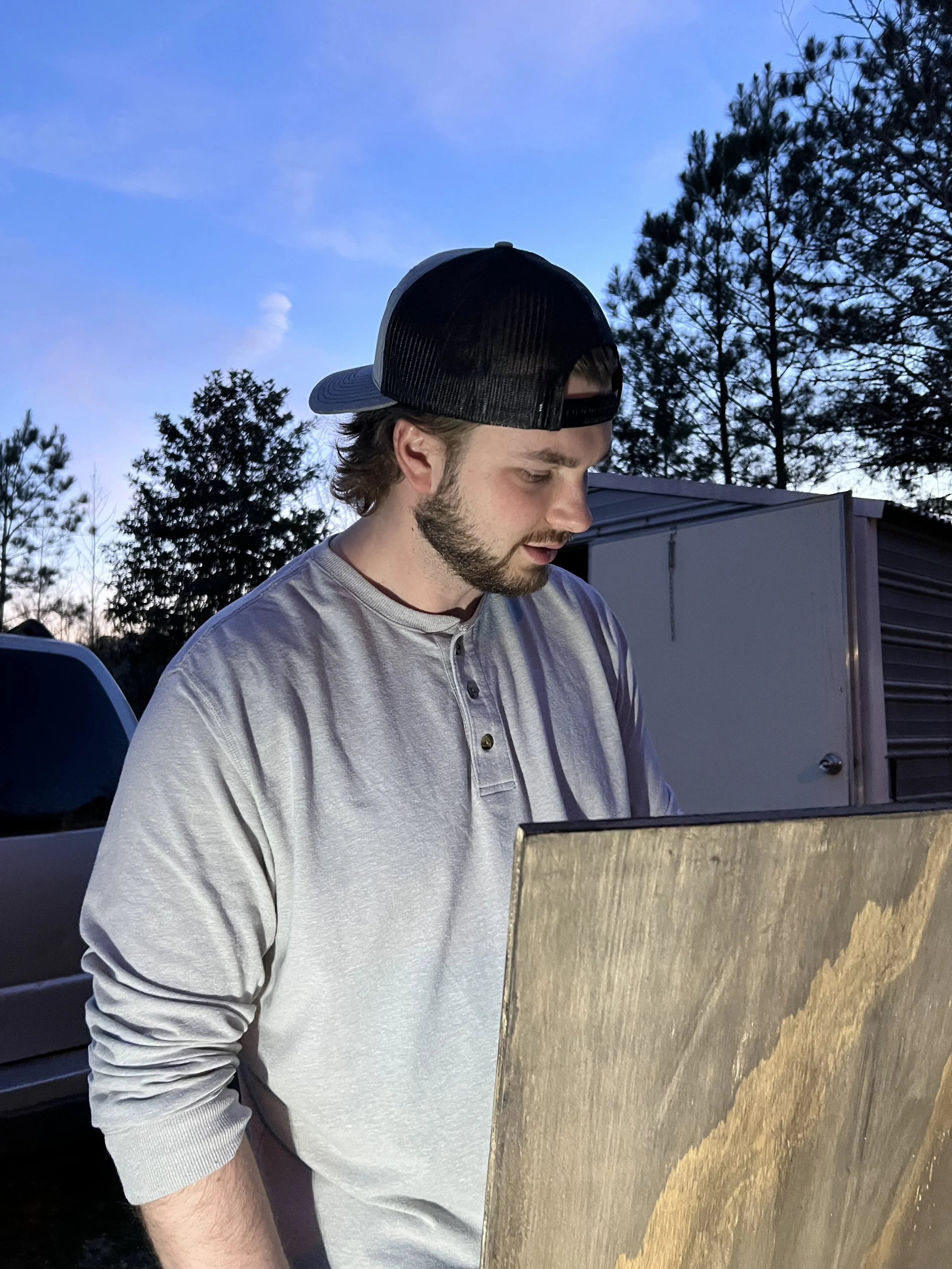 A young man with a beard wearing a black and white baseball cap, looking down, standing outdoors during twilight near parked vehicles and trees.
