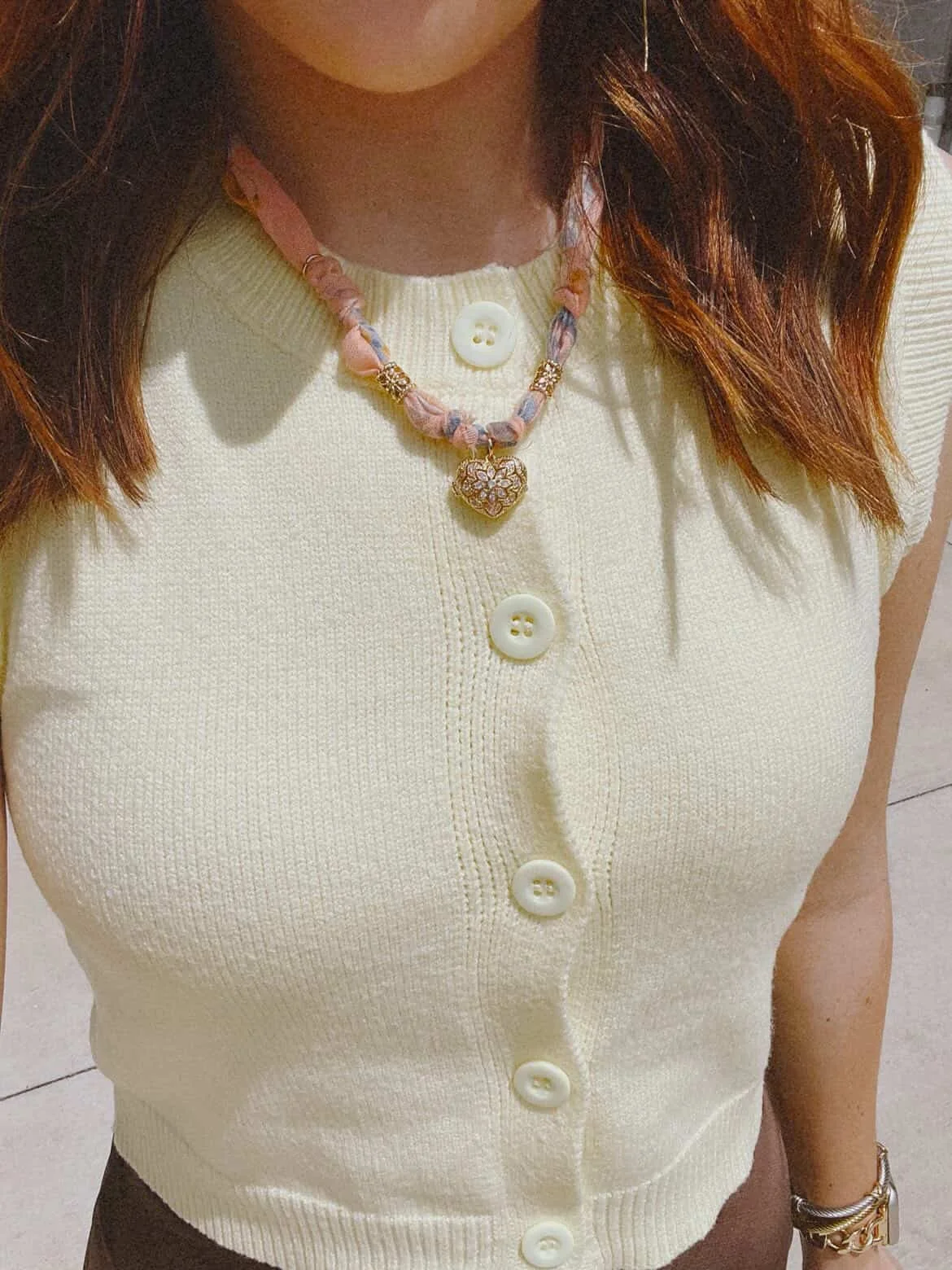 A woman wearing a yellow sleeveless button-up sweater with pearl buttons, a heart locket bandana necklace with a heart-shaped pendant, and a gold watch.