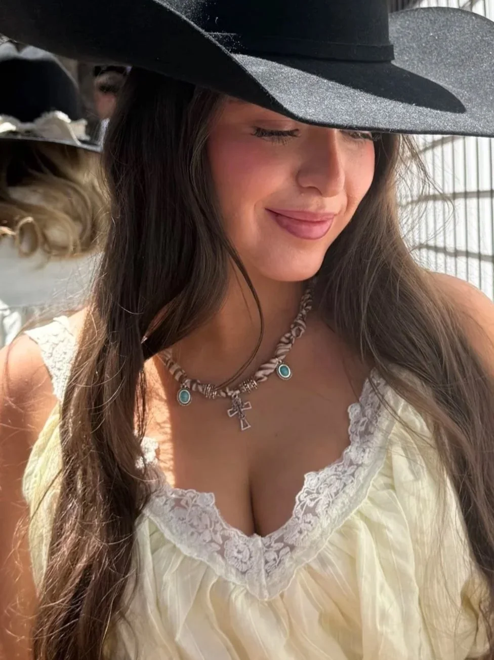 Woman with long dark hair wearing a large black cowboy hat, a white lace top, and bandana necklace that includes a cross necklace and turquoise earrings, smiling with eyes closed.