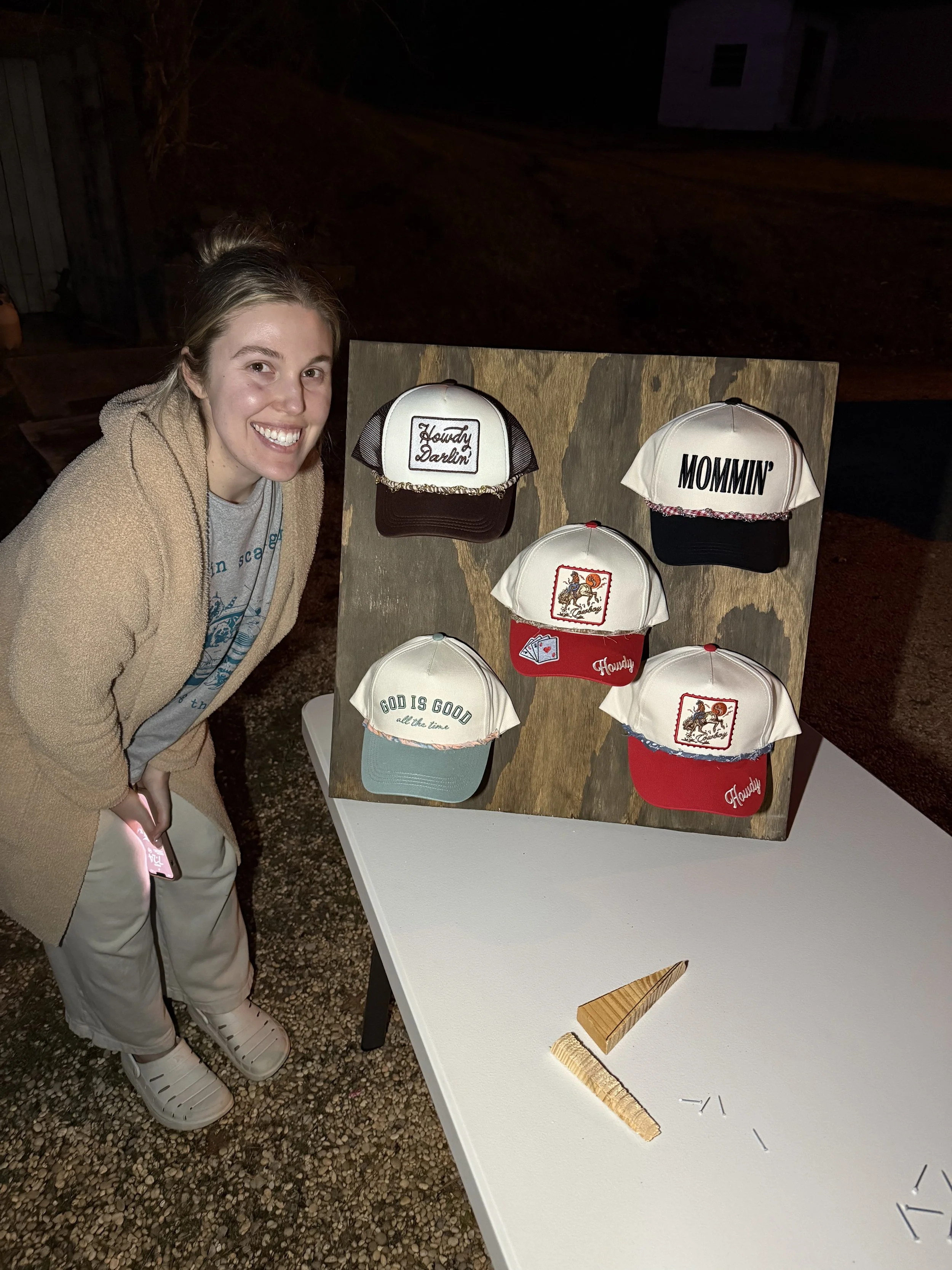 A woman smiling, dressed in a beige fleece jacket and khaki pants, standing next to a display of five baseball caps on a wooden board. The caps have various designs and sayings, including one that says 'Howdy Darlin'' and another that says 'MOMMIN'. The scene appears to be outdoors at night with a dark background and a white table beneath the display.