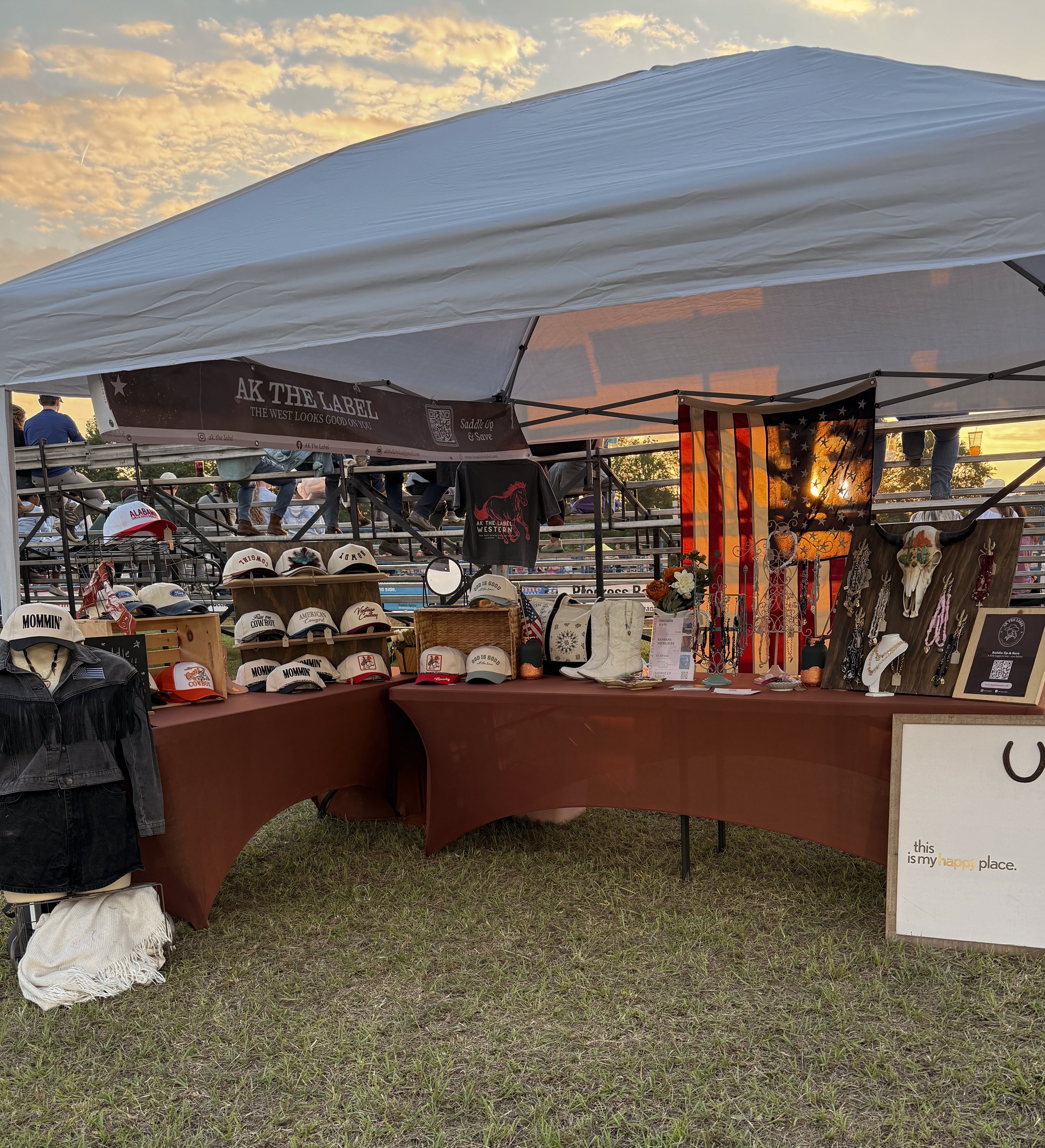 A ak the label booth at an outdoor pro rodeo with various necklaces, earrings, and decorative items on display, under a tent with a sunset sky in the background.