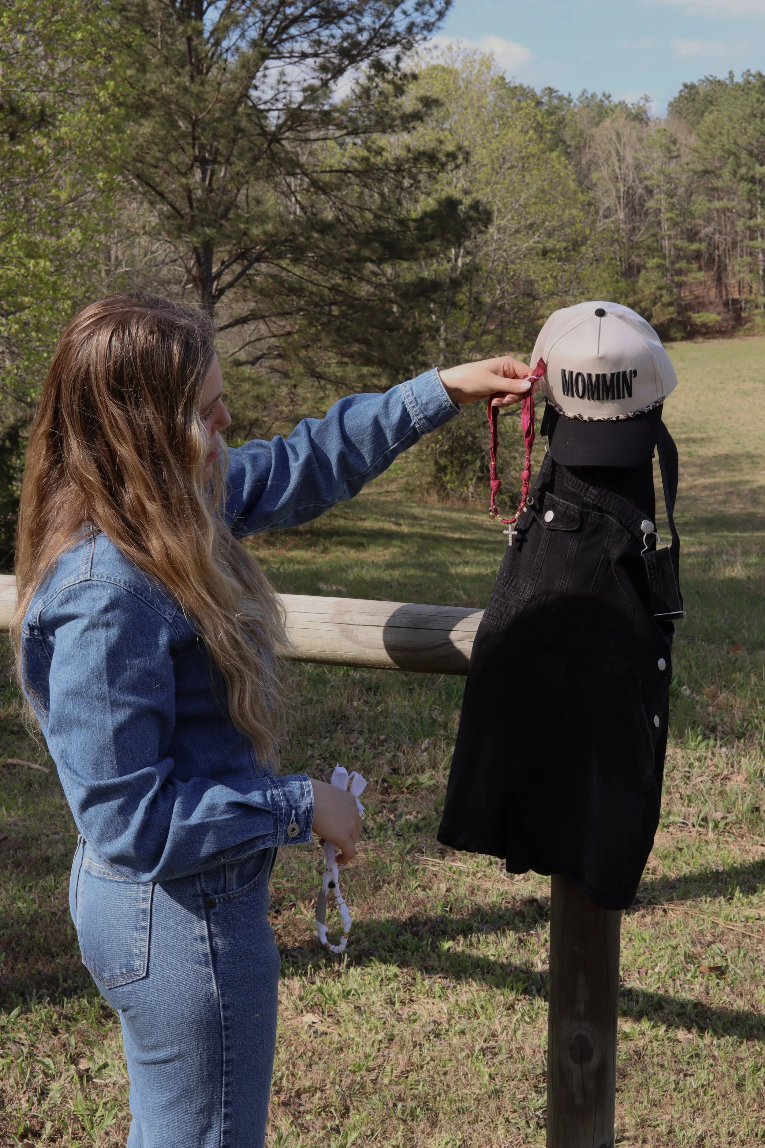 A young woman in a denim jacket and jeans holding a rosary stands next to a black backpack on a mannequin dressed in a white cap with the word "MOMMIN" embroidered on it. The scene is outdoors with trees and a grassy field in the background.
