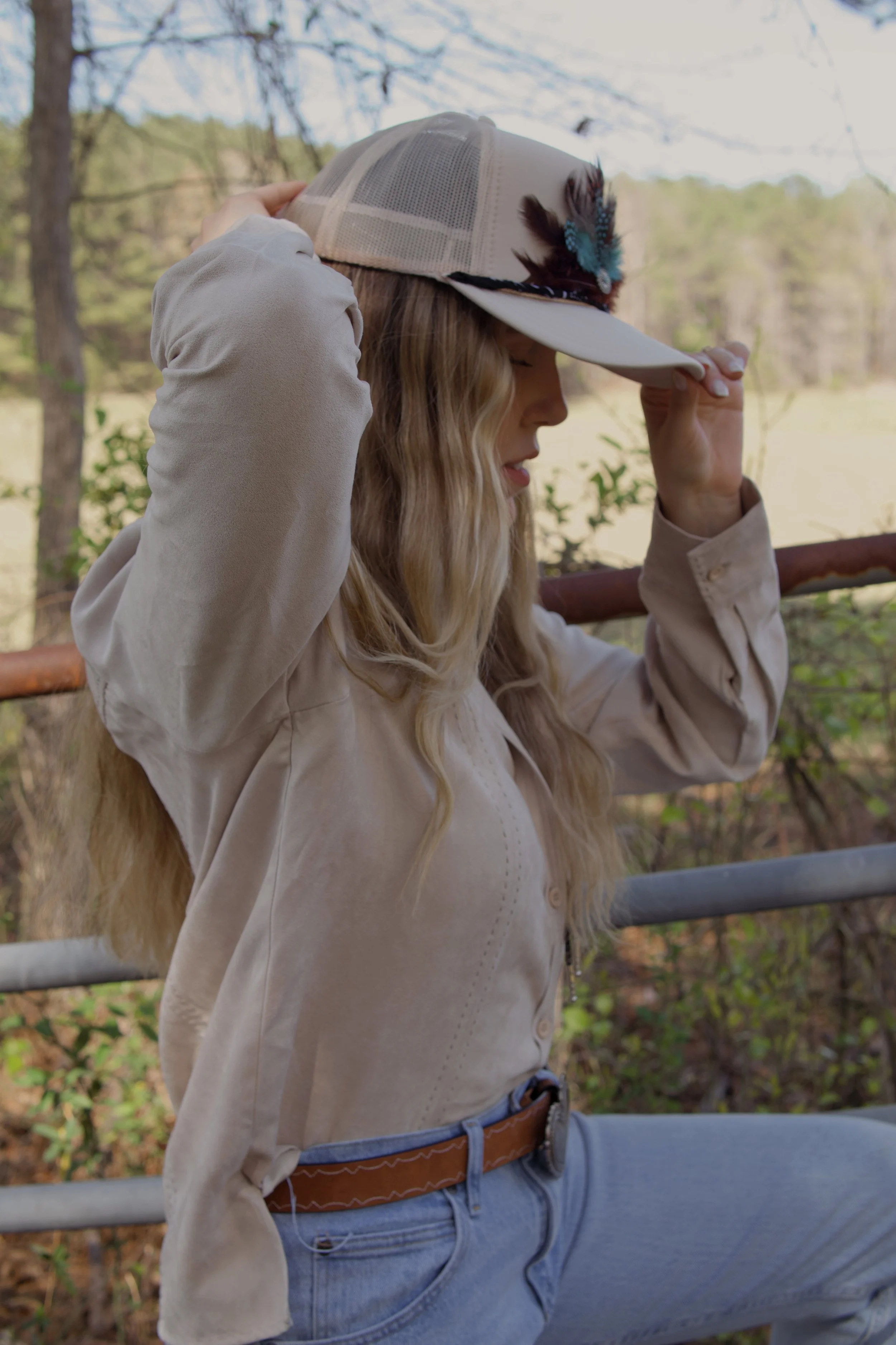 A woman with long blonde hair, wearing a beige jacket, blue jeans, and a tan belt, is adjusting a beige trucker hat with a feather decoration while standing outdoors near trees and a railing.