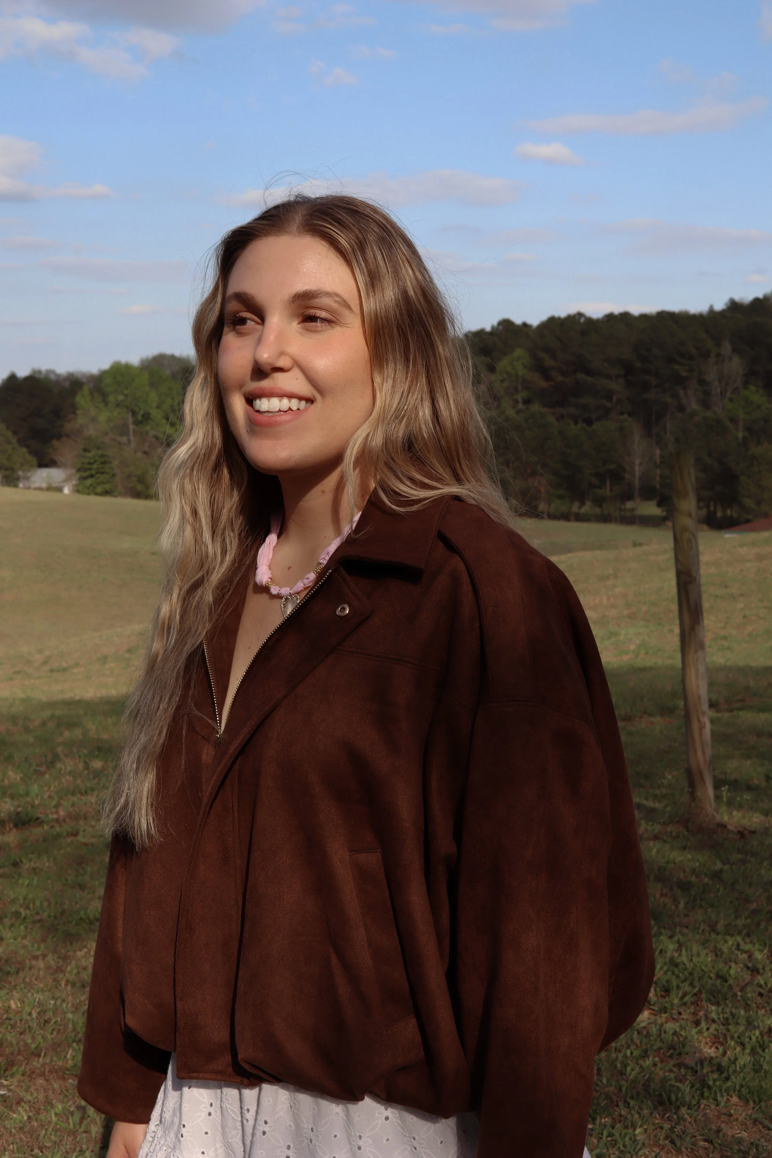 A young woman with long blonde hair smiling outdoors in a grassy field with a blue sky and trees in the background, wearing a brown jacket and white dress.