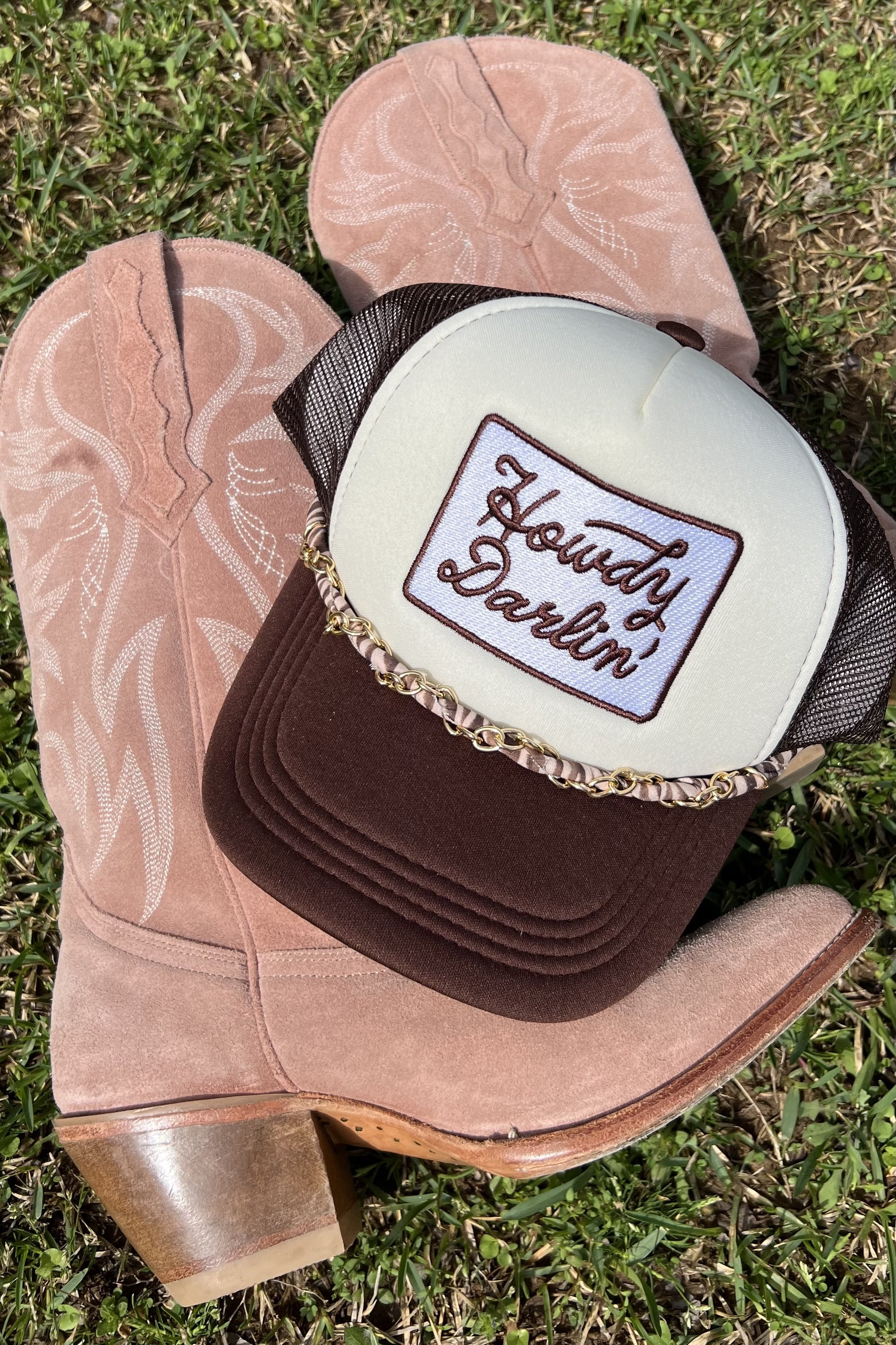 A cowgirl hat with a patch that says 'Howdy Darlin' on it, resting on a pink cowboy boot on grass.