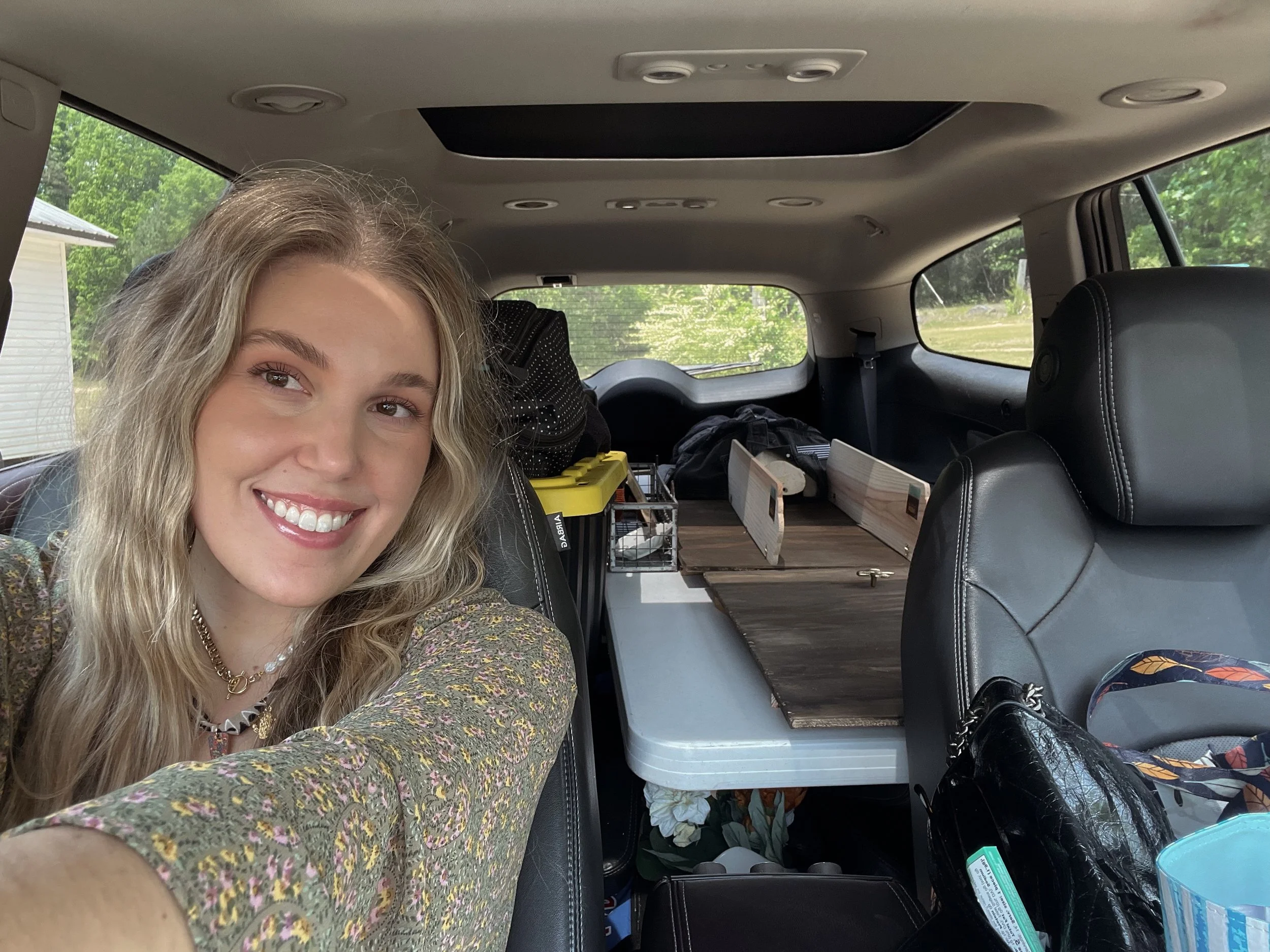A smiling woman with blonde curly hair takes a selfie inside a vehicle, with shelves and various items in the backseat and camper's interior visible.