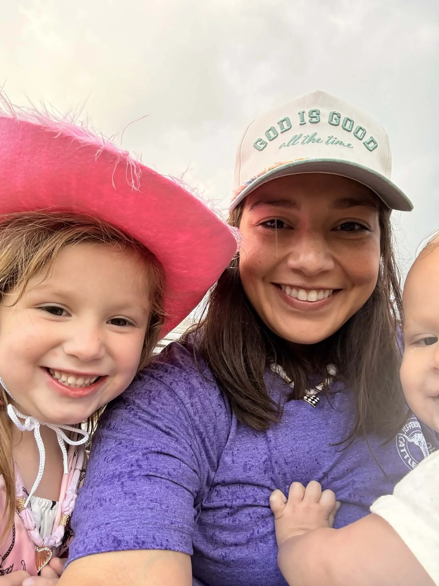 A smiling woman with long brown hair wearing a baseball cap that says 'Good is Good all the time.' She is taking a selfie outdoors with a young girl with red hair, wearing a pink hat and pink clothing. The sky is cloudy in the background.