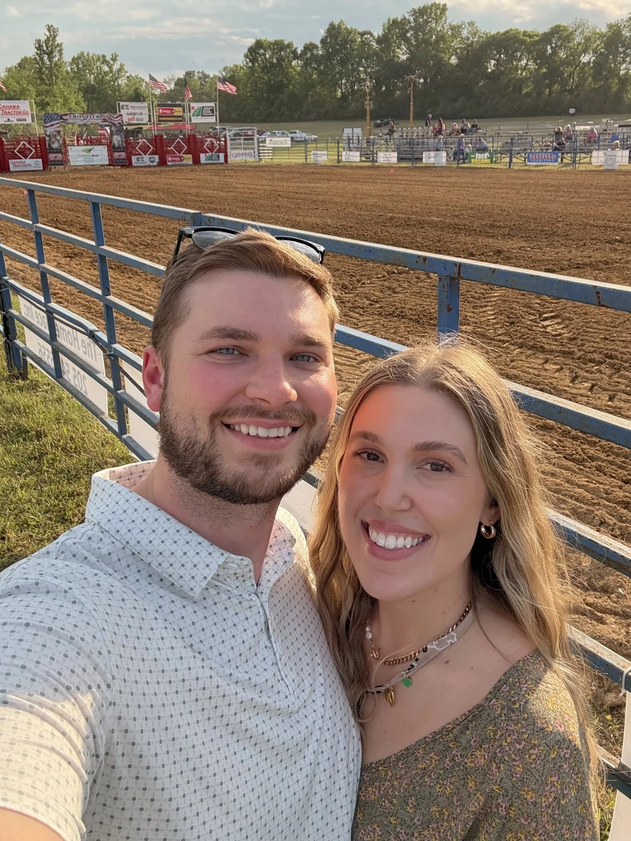 A smiling couple taking a selfie at a rodeo arena during the evening, with the dirt rodeo ground and spectator stands in the background.